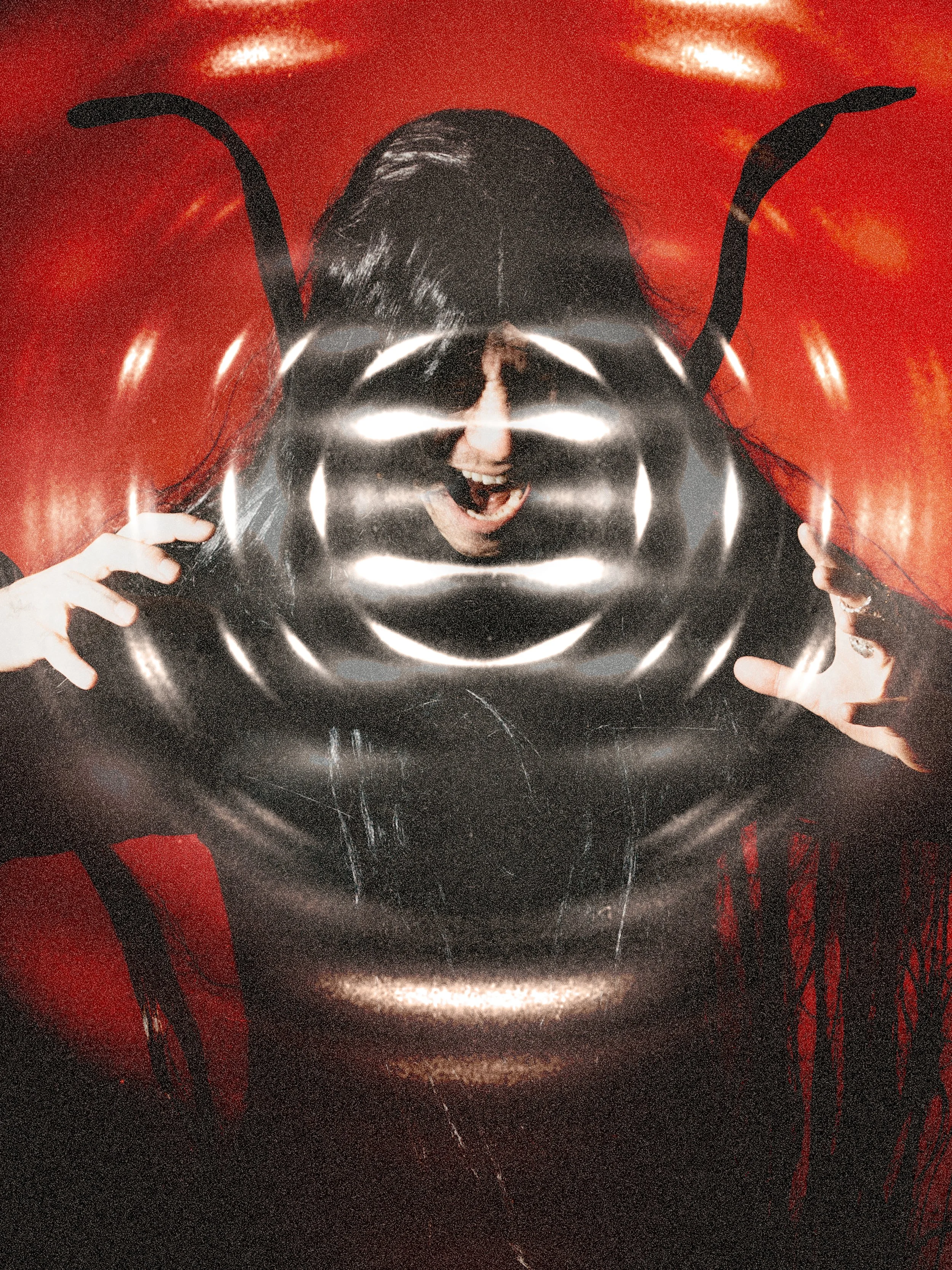 A woman with long black hair, open-mouthed, holding a black circular patterned object in front of her face, with red background and bright light reflections.