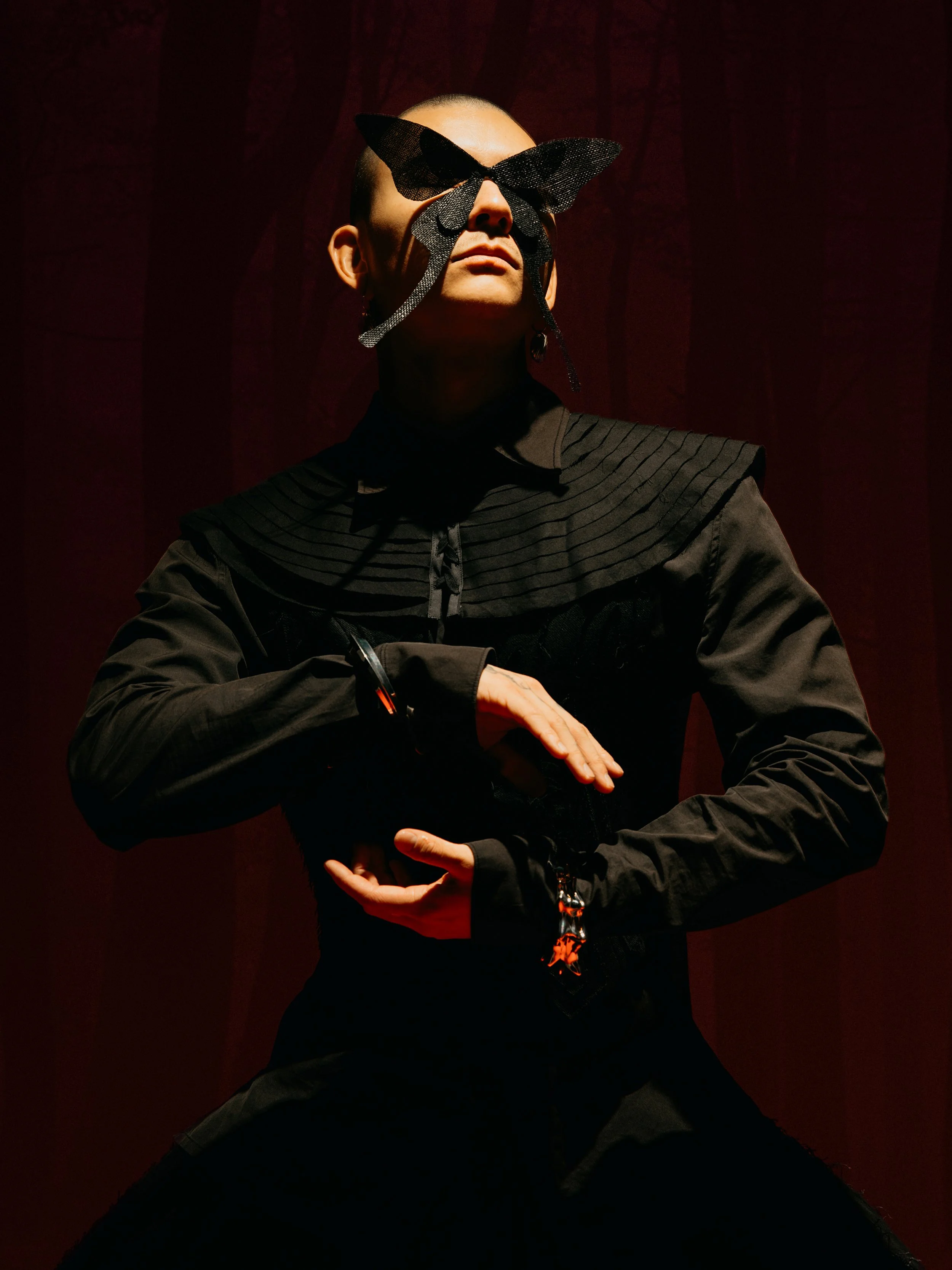 A person with a shaved head wearing geometric black sunglasses or a mask that covers his eyes. They are dressed in black clothing and posing against a dark red background with dramatic lighting.