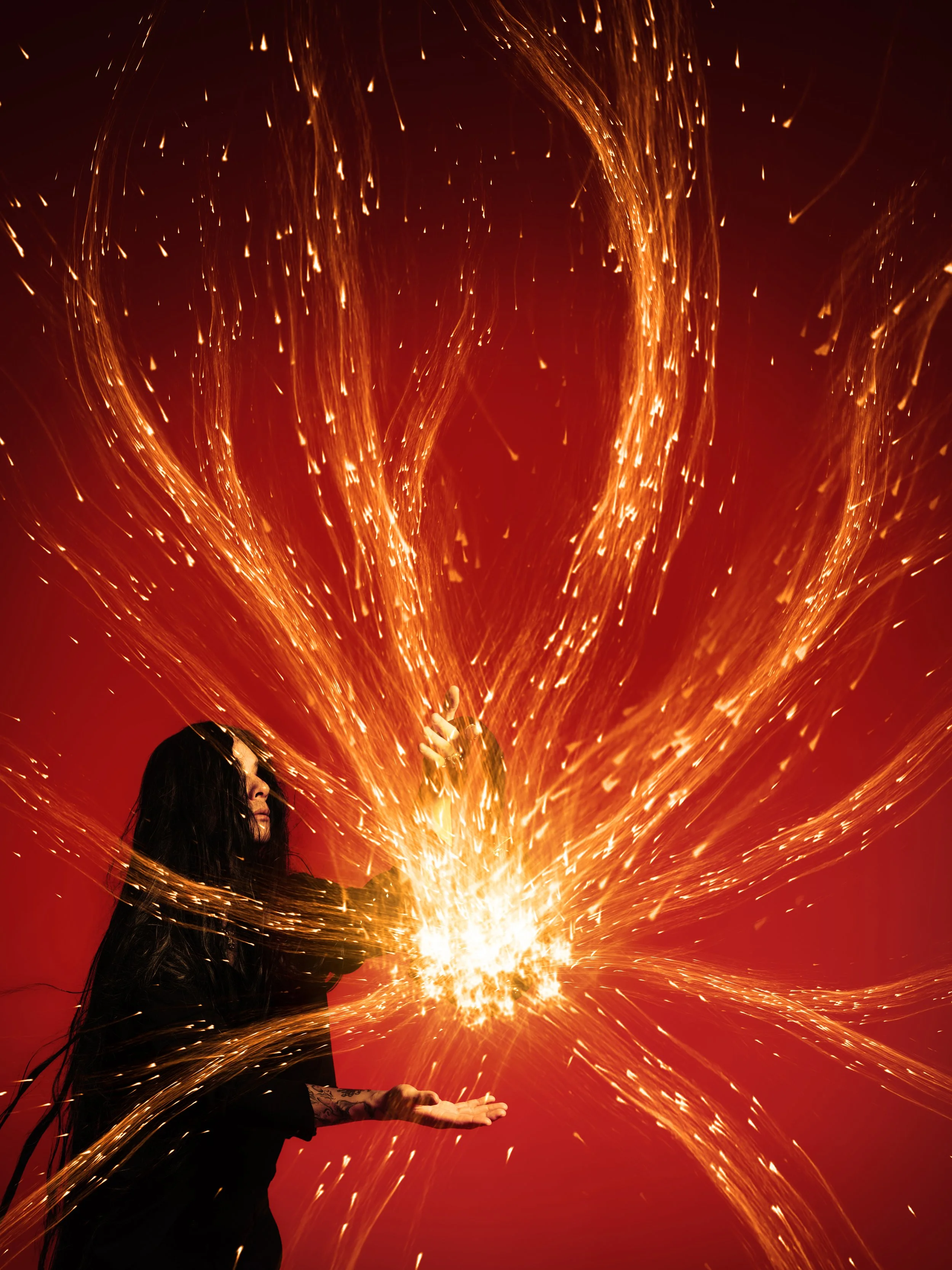 Person in black clothing with long dark hair holding a sparkler, with bright sparks flying in a dynamic pattern against a red background.