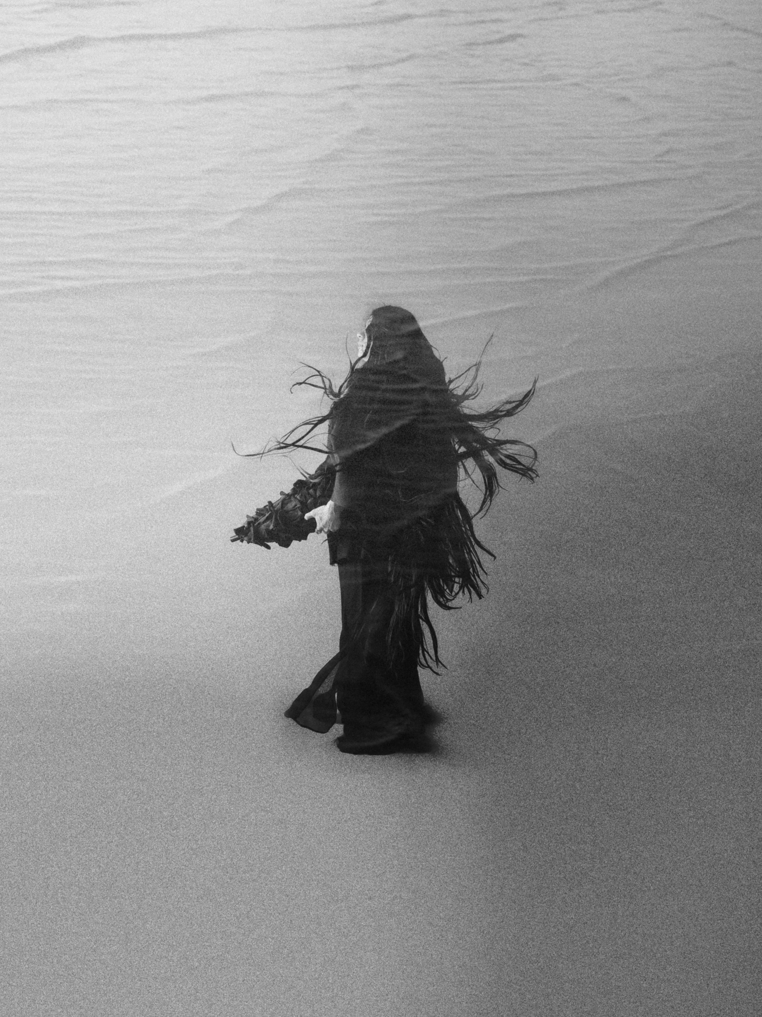 A person walking on a beach with long hair flowing in the wind, holding an object in their right hand, surrounded by smooth sand and gentle waves in the background.