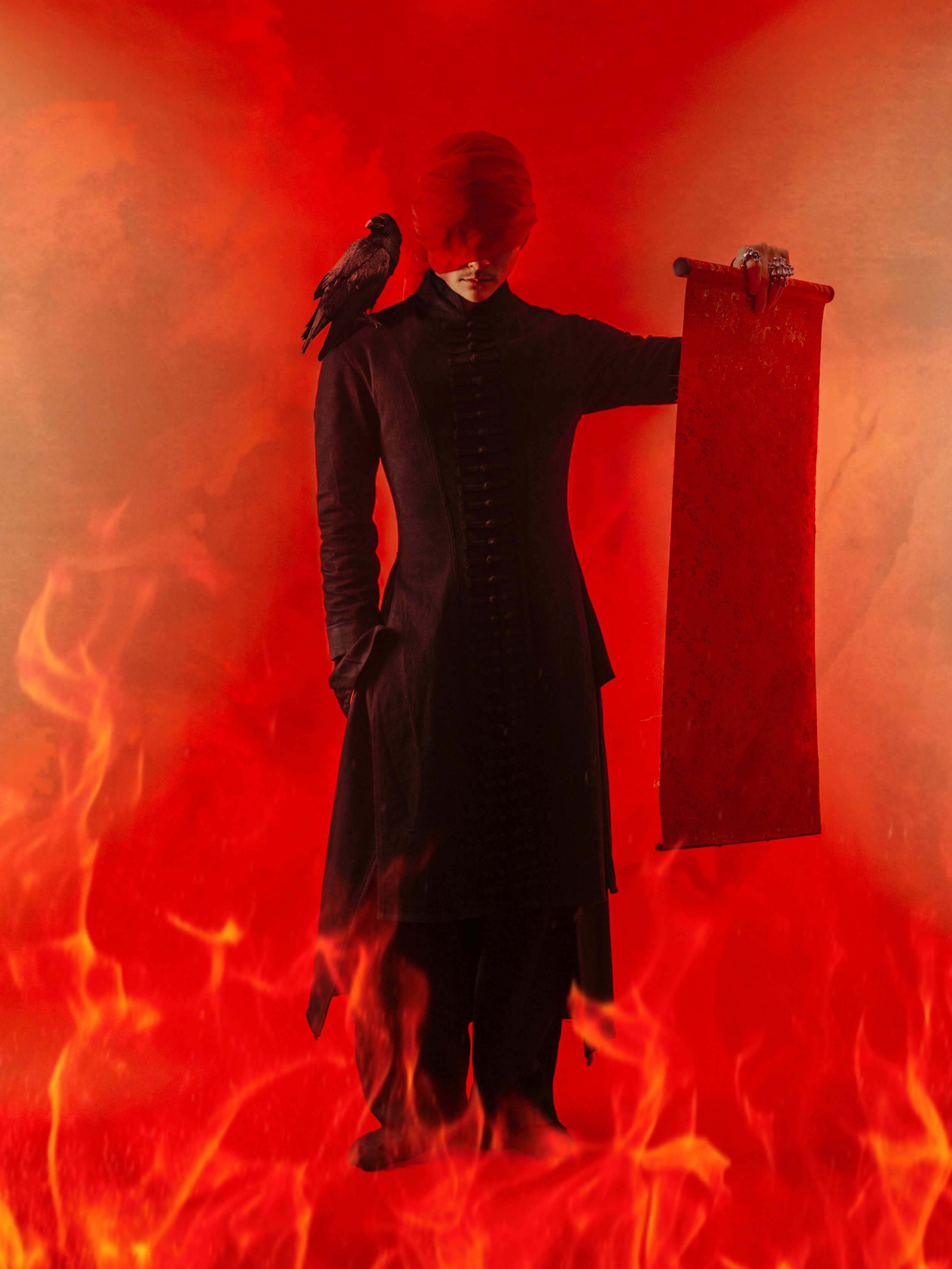 A person with a red head covering and black clothing stands in front of a fiery background, with a black bird perched on their shoulder and holding a red scroll.