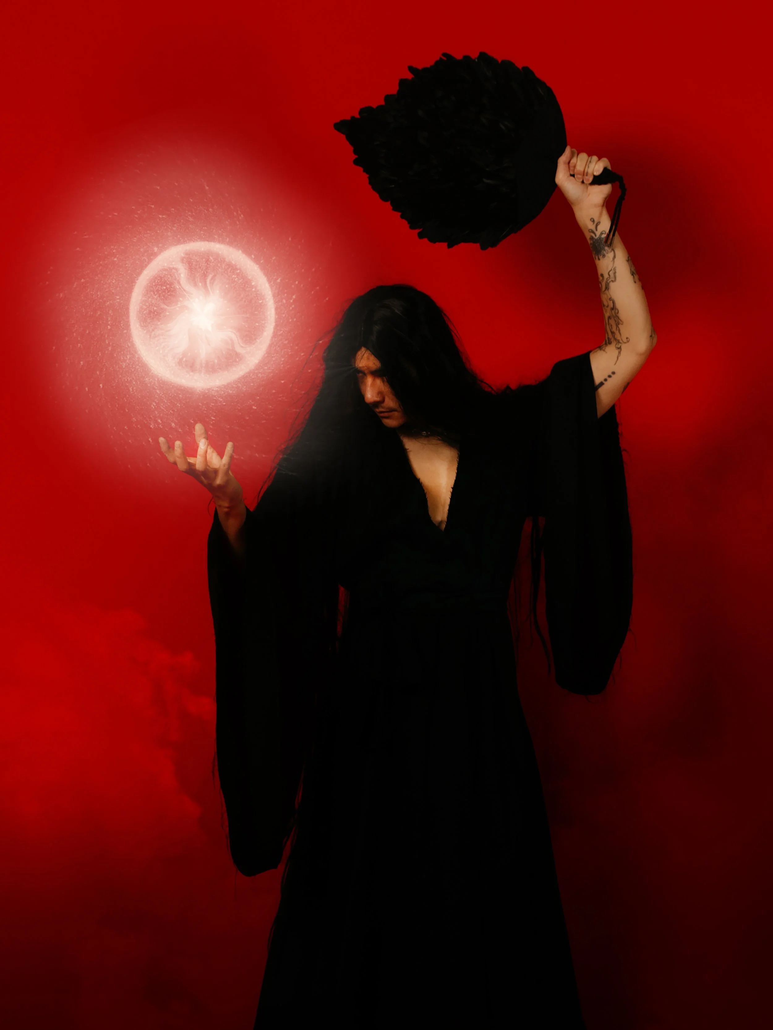 Woman with long black hair and tattoos holding a black feathered fan in front of a red background, with a glowing white circular pattern near her hand.