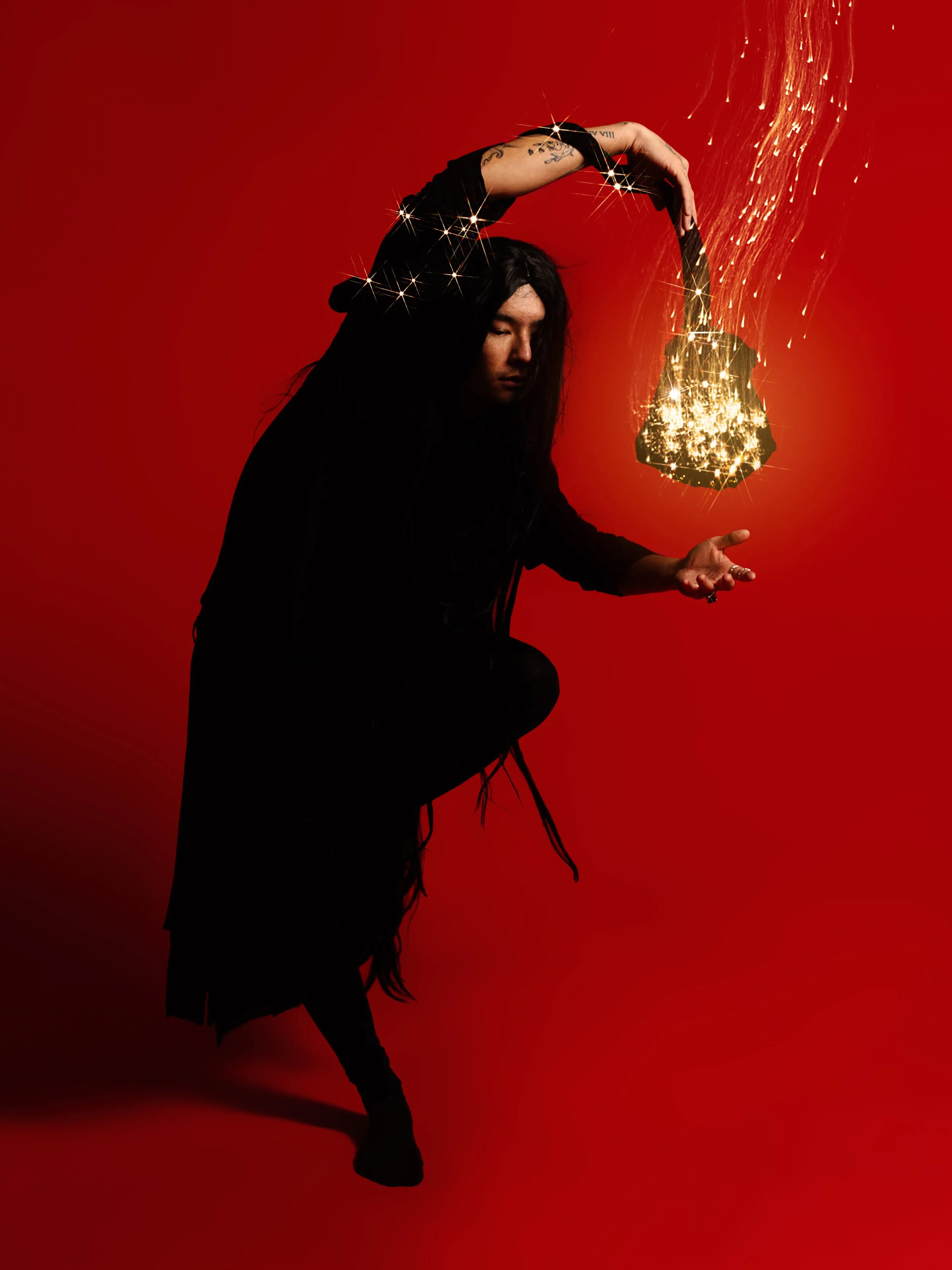 A person with long dark hair, tattoos on the arm, and dressed in dark clothing, is holding a sparkling object that resembles a rock or gemstone with sparks, against a red background.