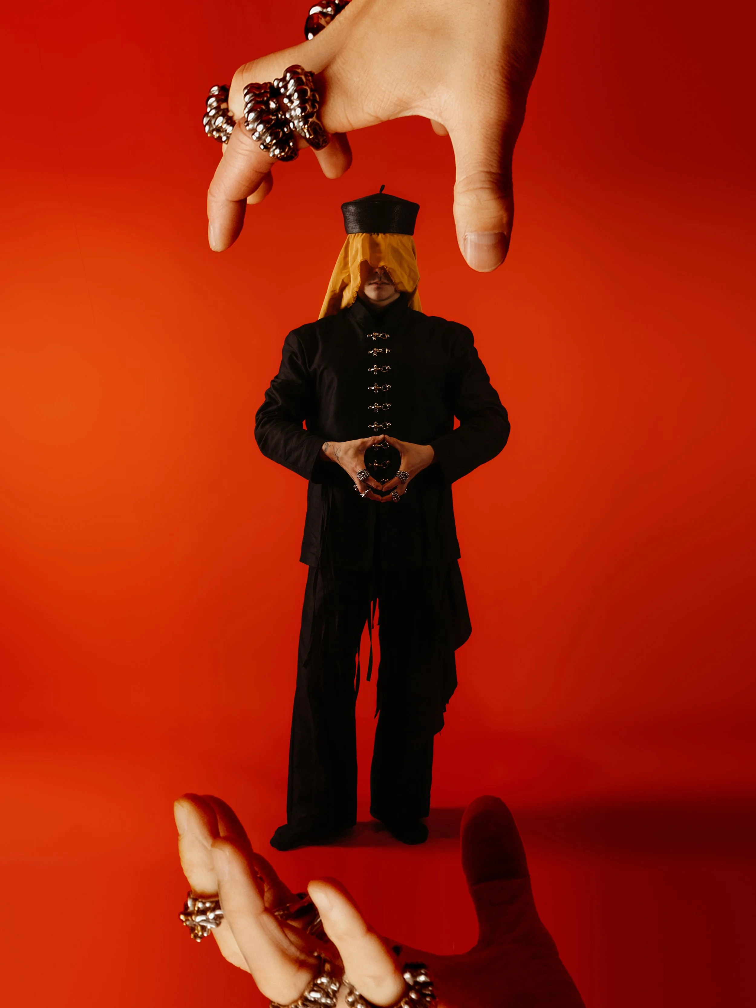 A person dressed in black with a yellow head covering and multiple rings, holding a miniature figure of themselves against a red background. The image is reflected in a mirror, creating a symmetrical effect.