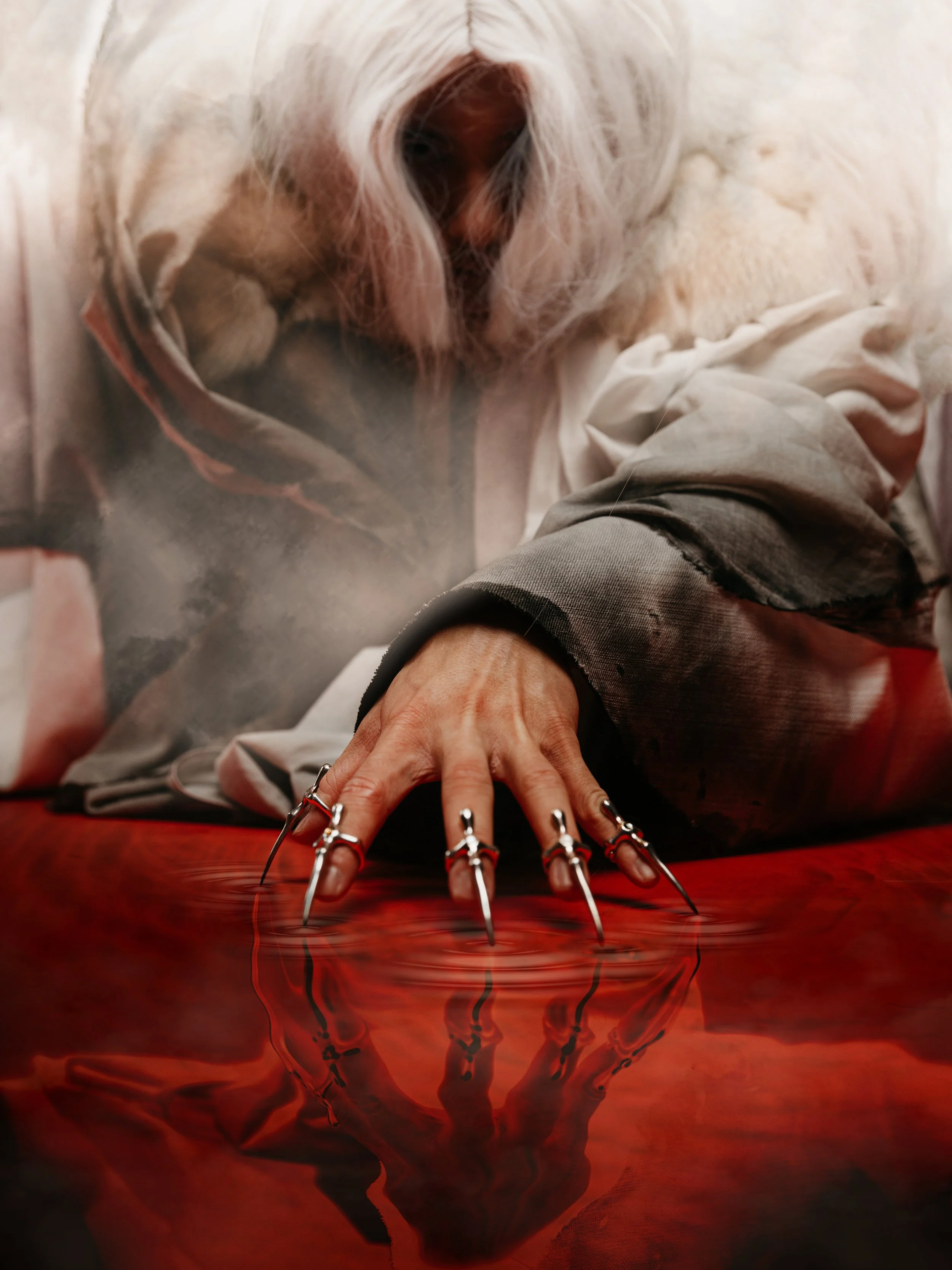 A person with long nails and rings on their fingers is reaching towards a reflective surface with a red hue. The reflection creates a dark, animalistic shape beneath the hand.