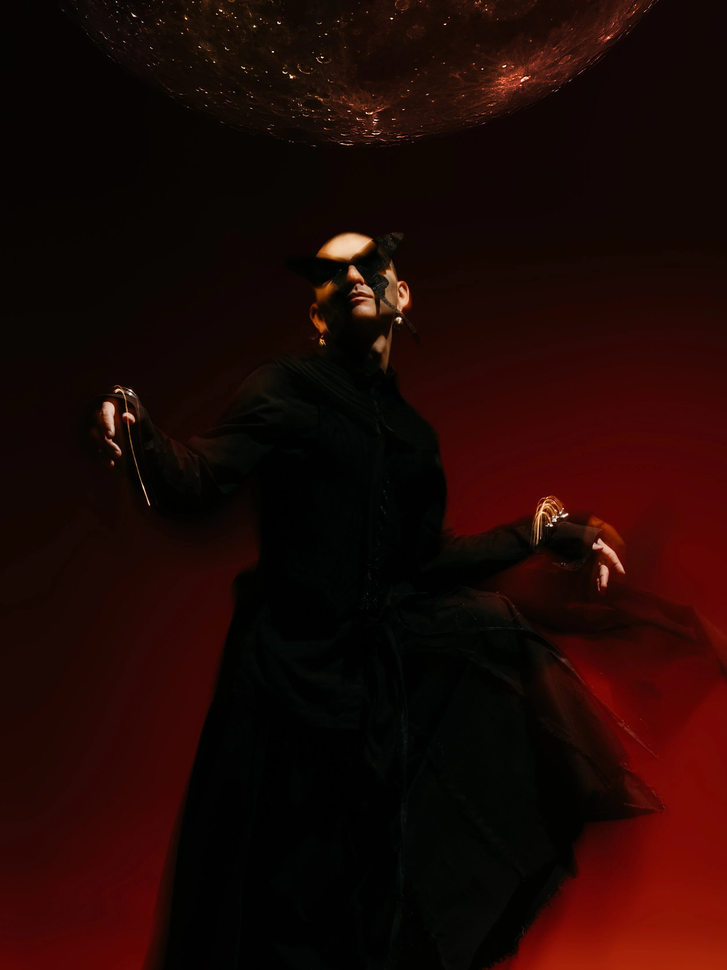 A person wearing a black jacket and black mesh mask with cat ears stands against a dark background with red lighting. The person has their arms outstretched and appears to be looking at the camera.