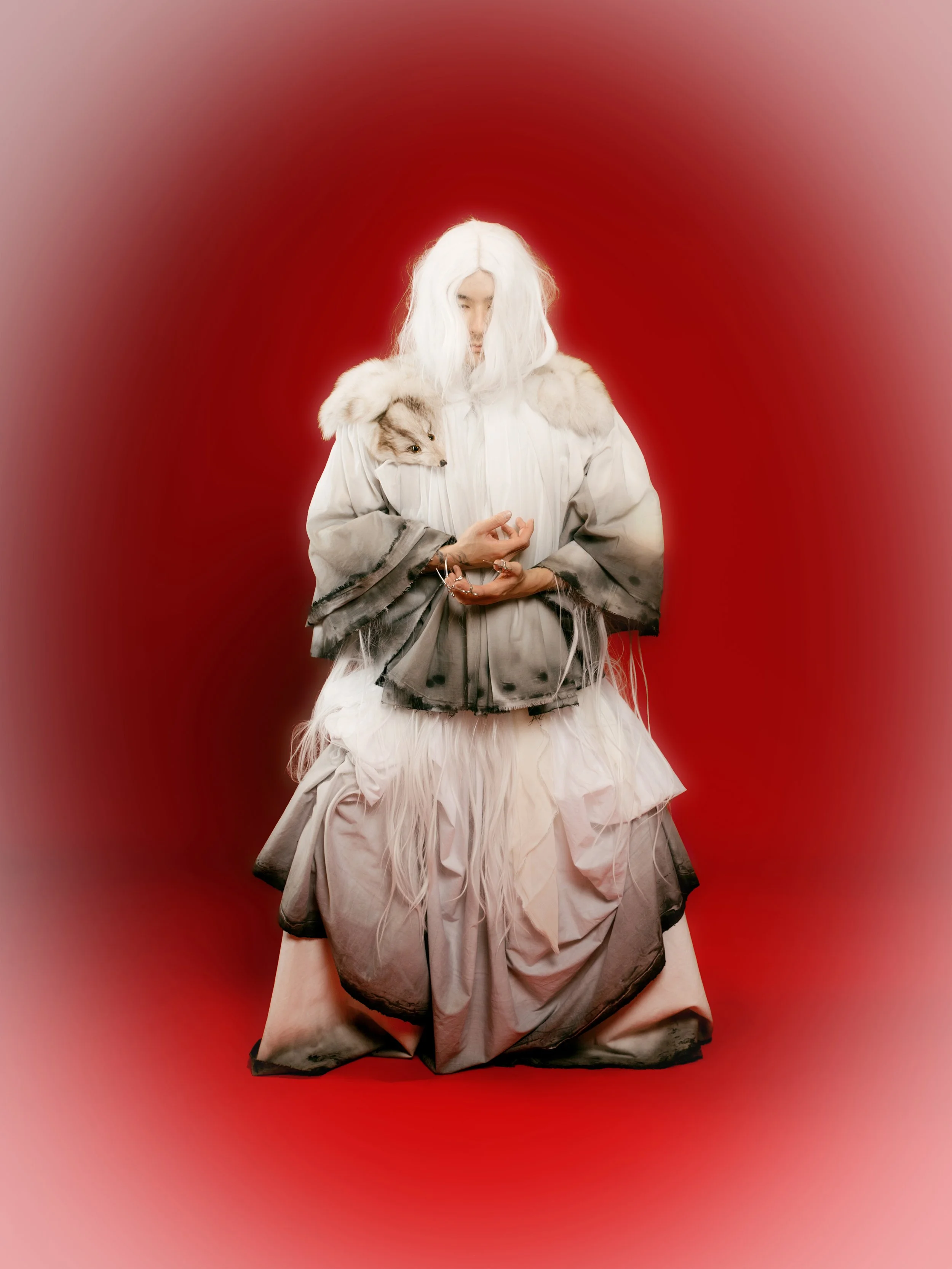 Person with long white hair wearing an elaborate dress with layers and a faux fox fur shawl, standing against a red background.
