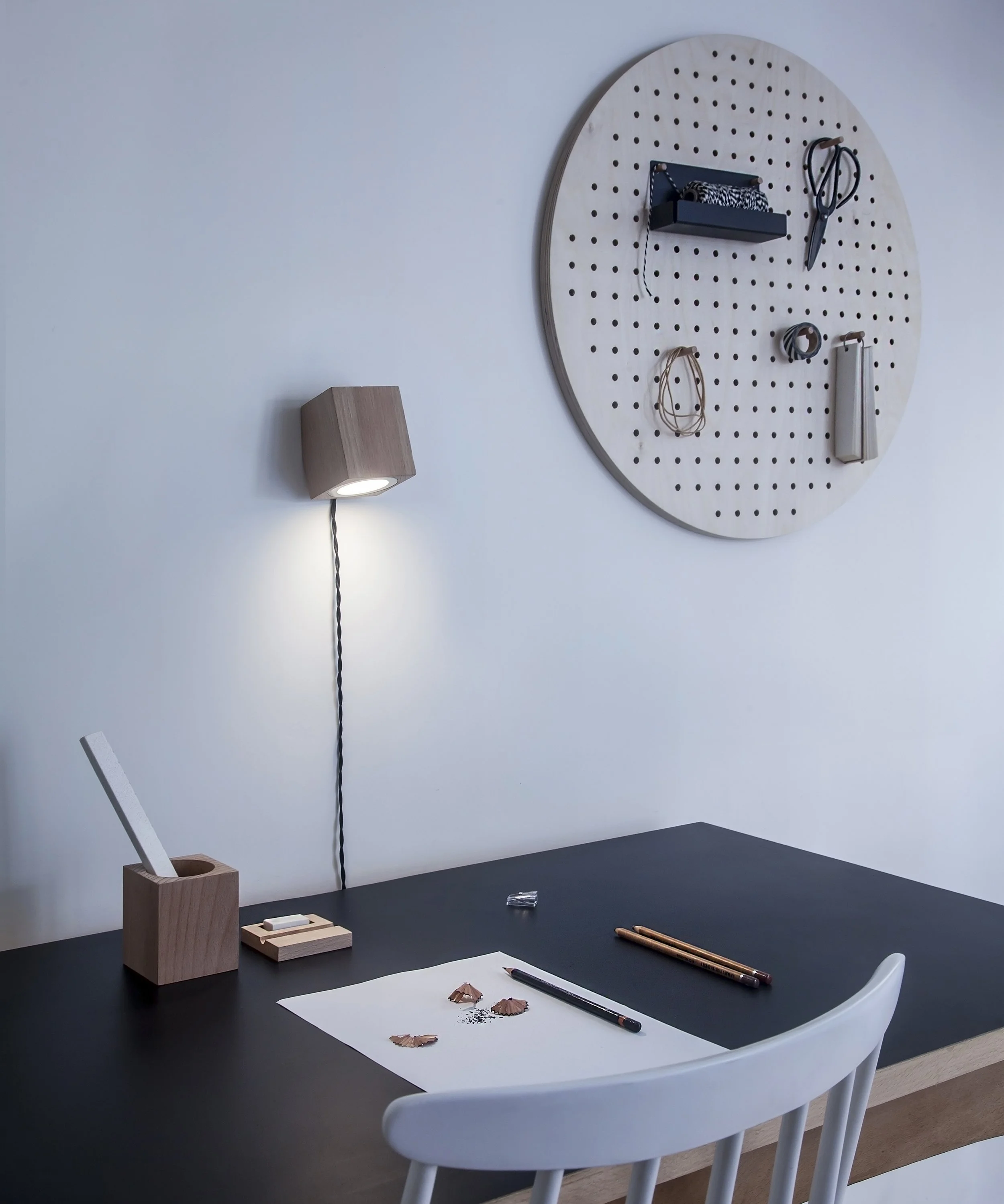 A modern workspace with a black desk, a white chair, and a cork tissue box. There is a wall-mounted light, a pegboard with various office supplies, and a piece of paper with torn paper edges, pens, and a highlighter.