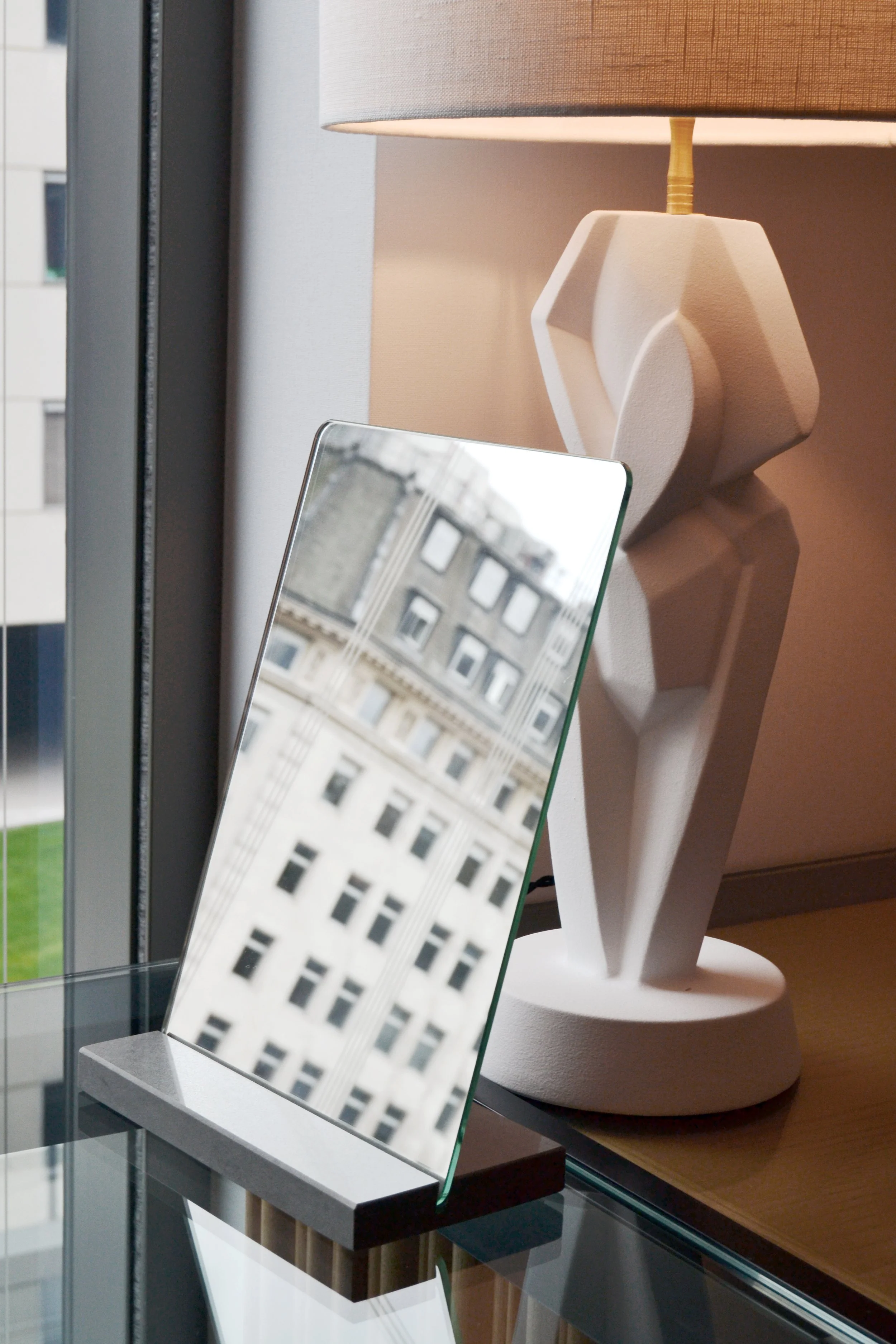 Decorative table lamp with abstract white base and beige shade, next to a tilted mirror reflecting a building exterior, on a glass-topped table.