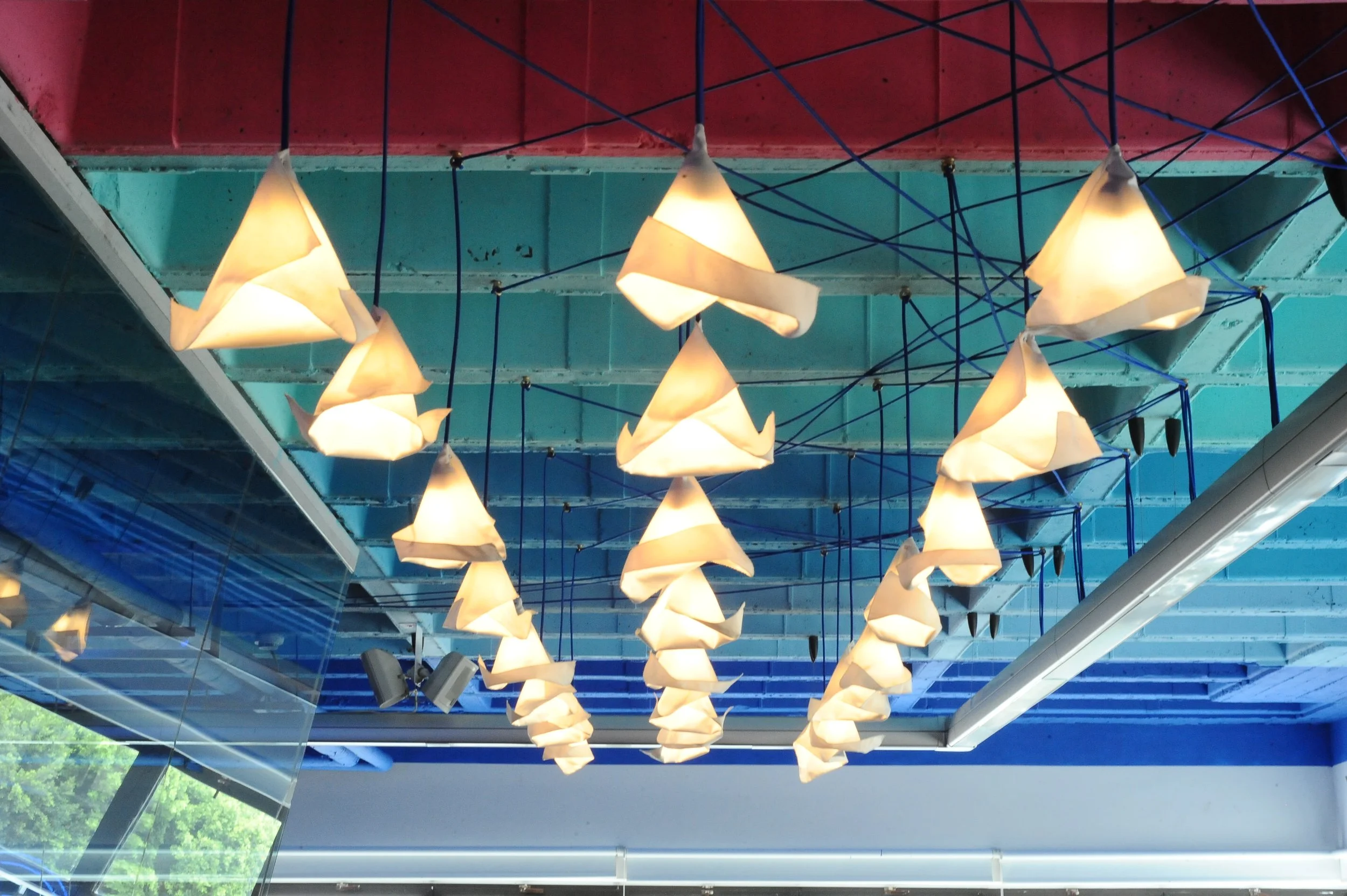 Ceiling lights shaped like paper cones hanging from blue and red cables in a modern indoor space with glass wall and green trees outside.