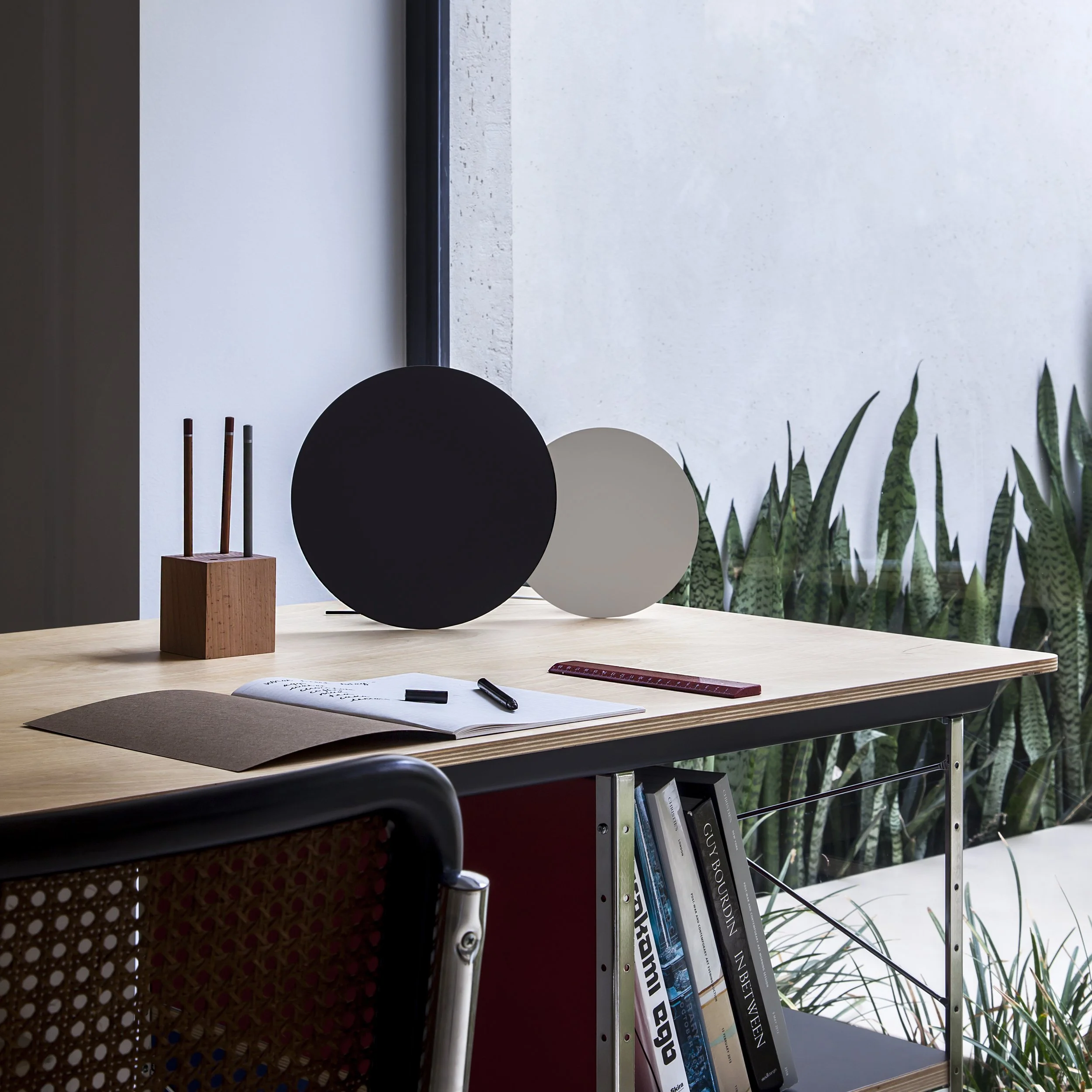 Modern workspace with a light wood desk, two circular decorative objects, a ruler, an open notebook with pens, and a bookshelf with books underneath a large window with lush plants outside.