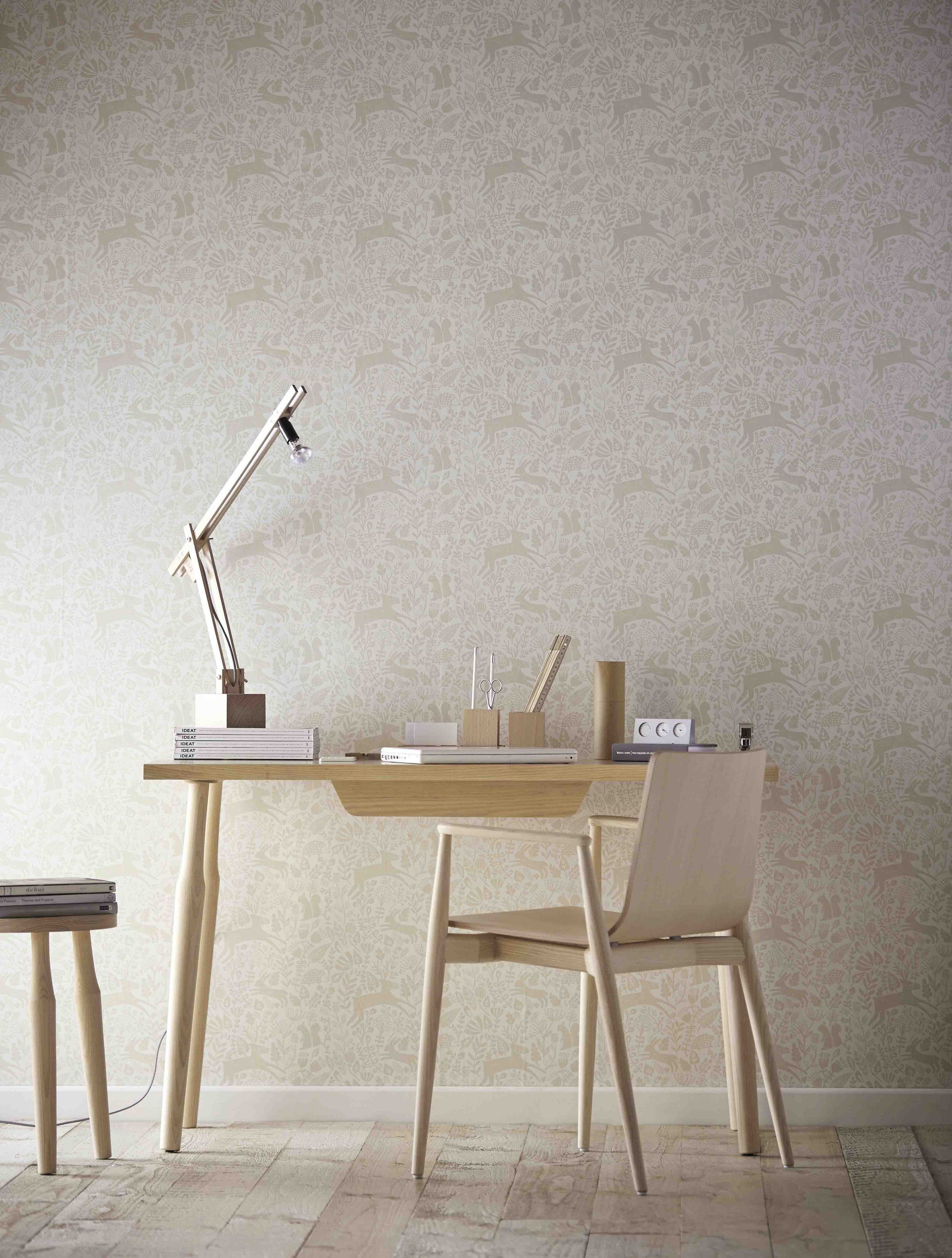 A minimalist desk setup with a wooden chair, desk, and accessories such as books, a lamp, and stationery, against a patterned wallpaper with woodland animals.