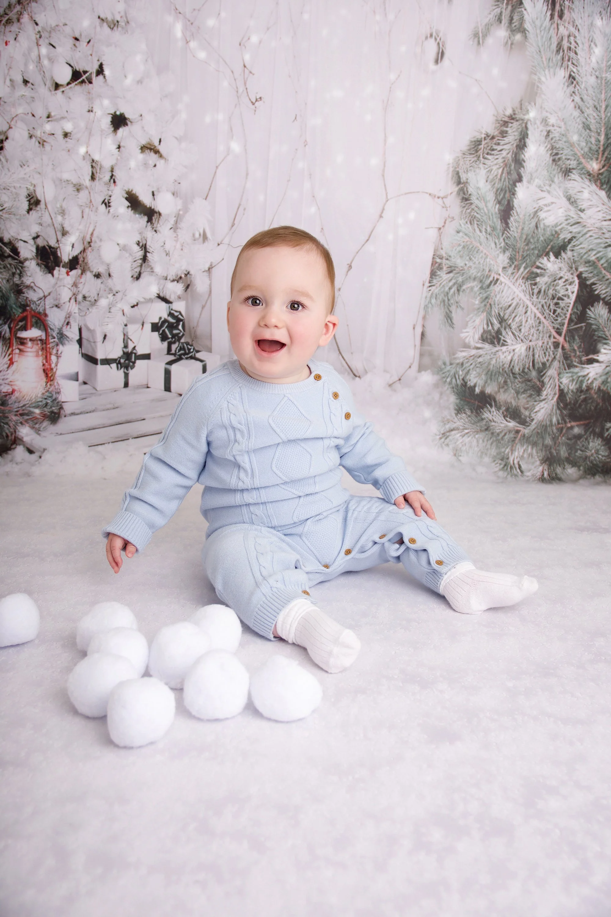 A smiling baby wearing a light blue sweater and pants, sitting on a snowy surface surrounded by white snow balls, with a wintery background of white trees and green pine branches.