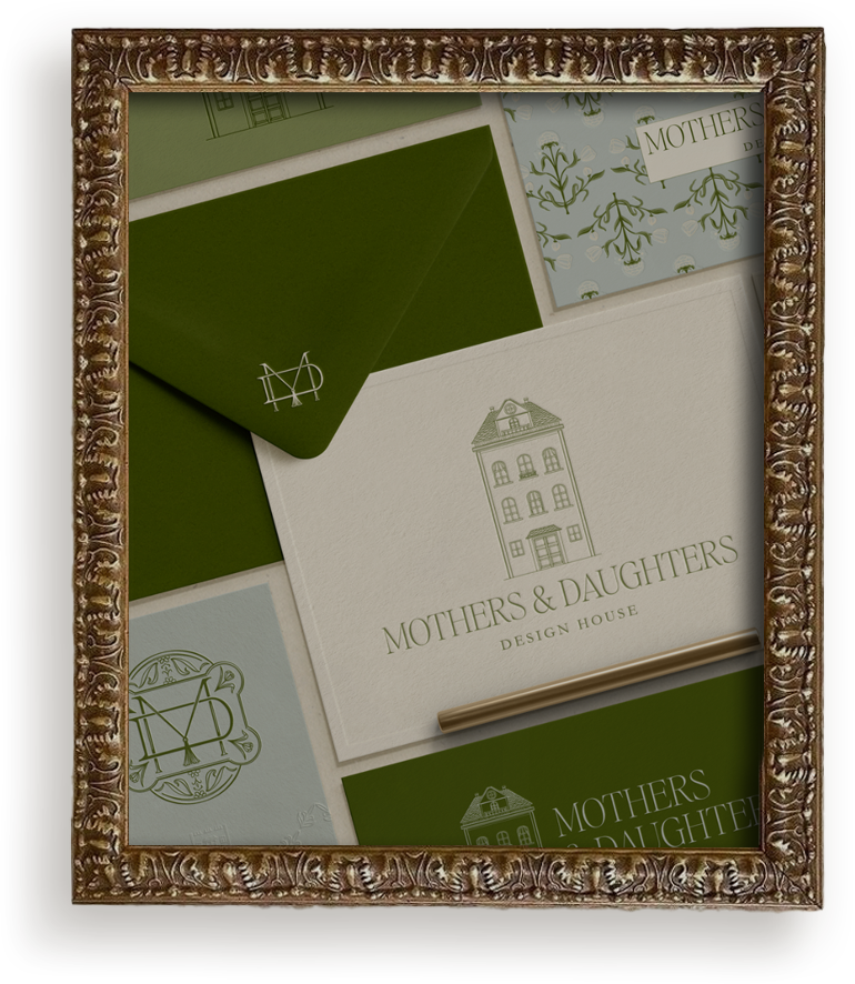 A display case containing various stationery and packaging samples from Mothers & Daughters Design House, including a white card with a house illustration and the company name, green envelopes, and patterned paper, all framed with an ornate gold frame.