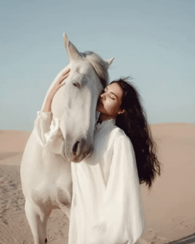 A woman hugging a white horse in a desert landscape under a clear sky.