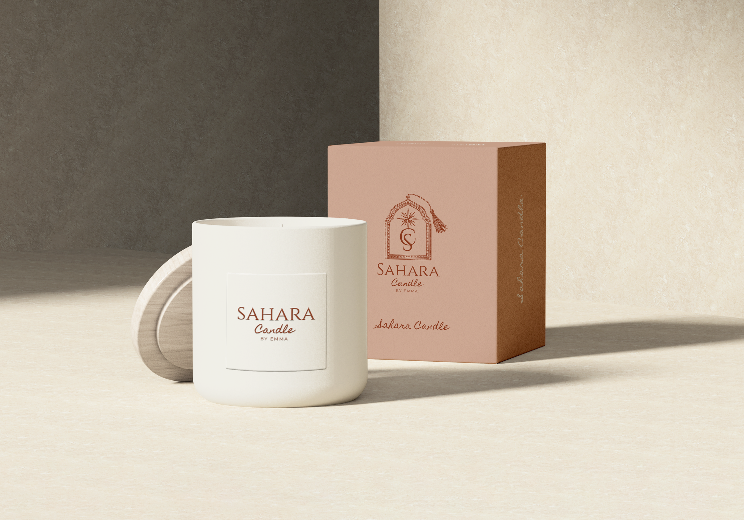 A white candle in a white holder with a label reading 'SAHARA Candle BY EMMA', next to a pink box with the same label and design, on a neutral surface with a shadowed background.