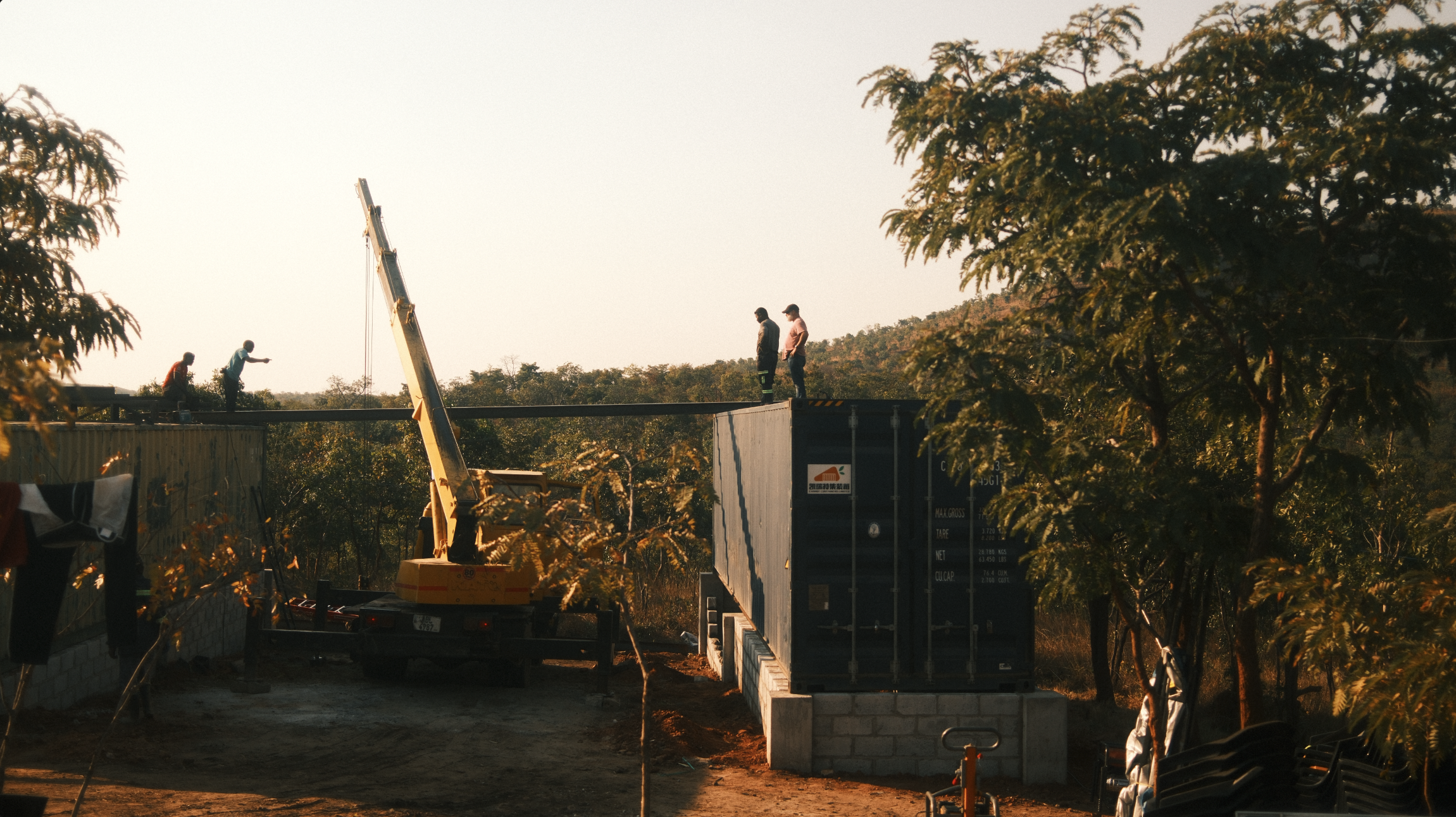 Construction workers standing on shipping containers beside a crane at a wooded outdoor worksite