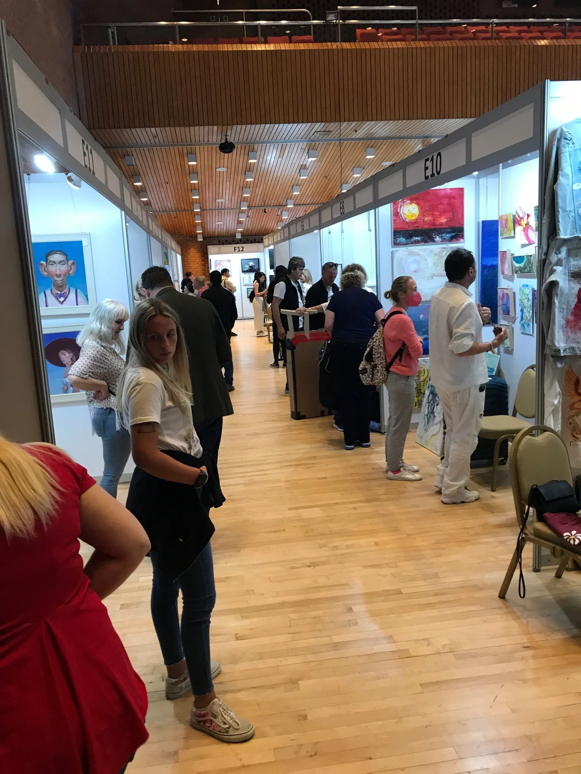 People browsing artwork at an art exhibition in a gallery hall with wooden flooring and booths labeled E10, E11, E12, E13, E14, E15.