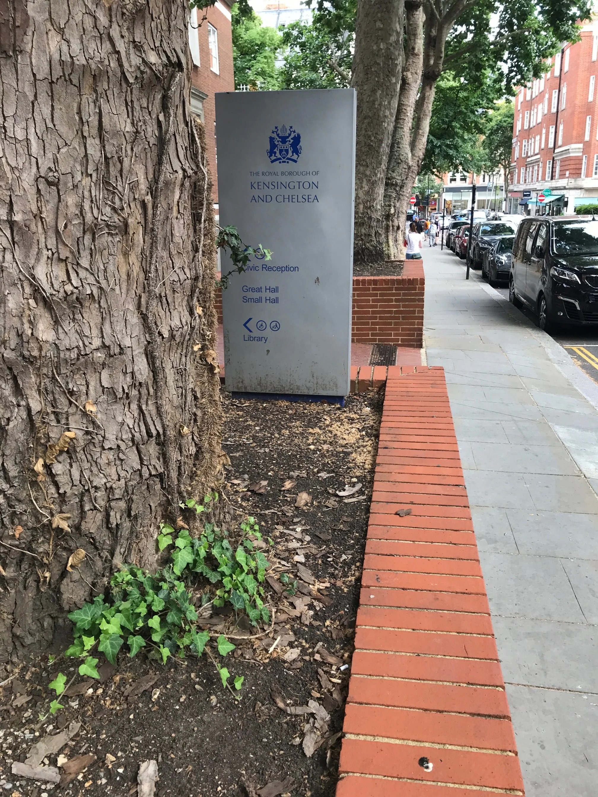 Sign for The Royal Borough of Kensington and Chelsea with directions to Civic Reception, Great Hall, Small Hall, and Library on a city sidewalk, with a large tree, parked cars, and brick buildings in the background.