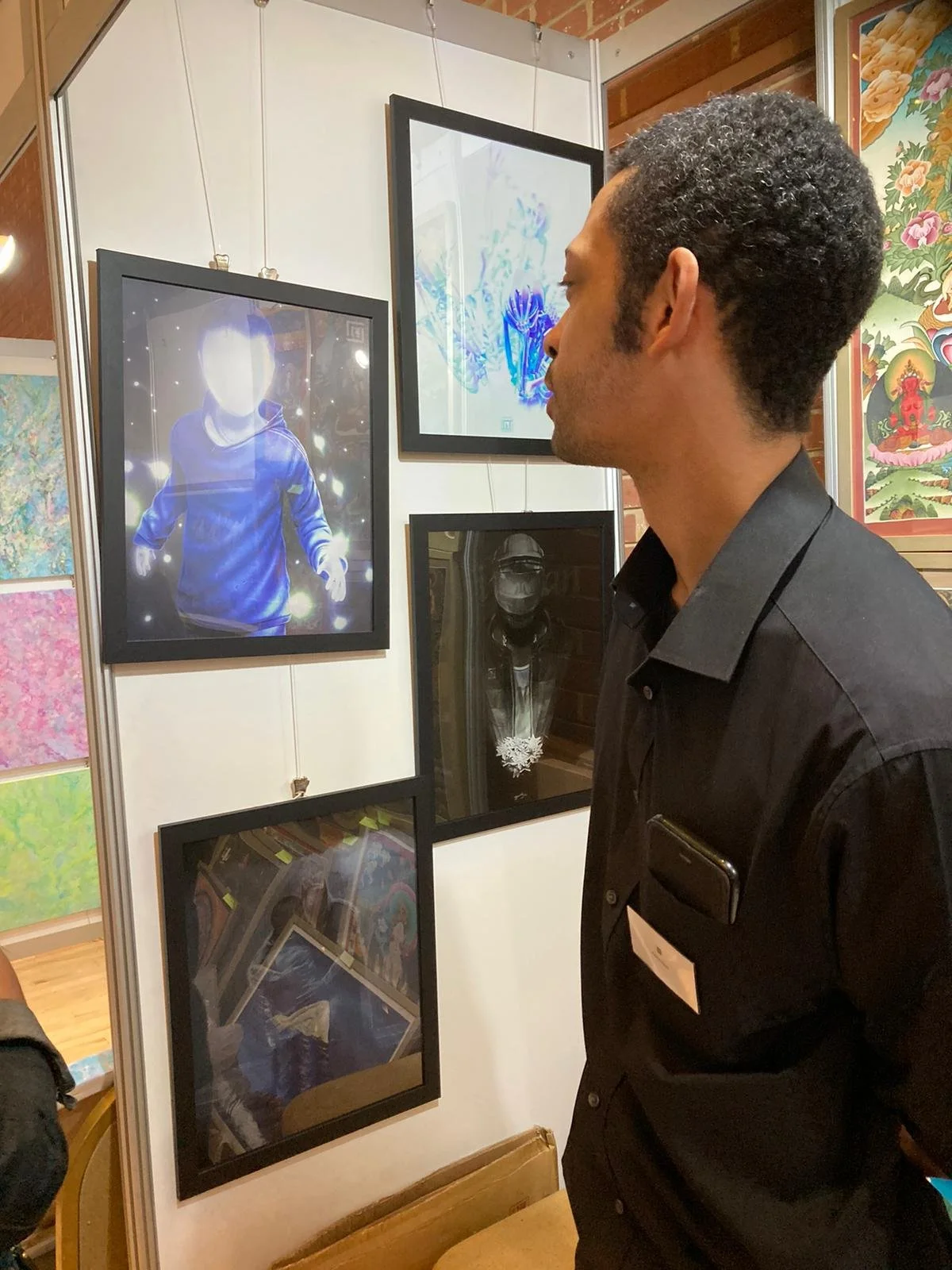 A man with dark curly hair wearing a black shirt and a name tag looks at framed artwork and photographs displayed on a white wall at an art gallery.