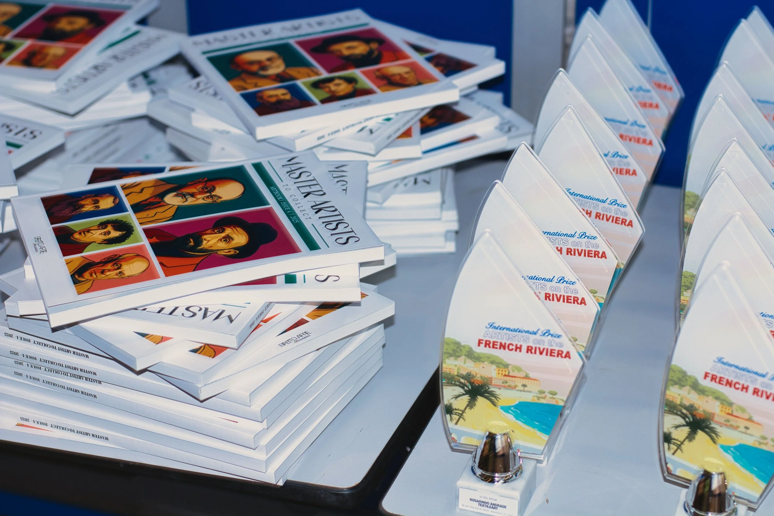 Piles of catalogs and brochures with artwork of famous painters on the covers, and several glass awards shaped like waves with colorful beach scenes and text indicating an international art contest in French Riviera.