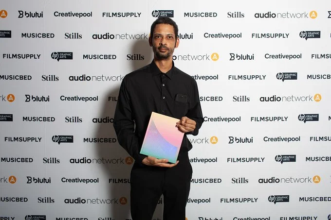 Man standing in front of a step and repeat backdrop, holding a tablet device, dressed in black.