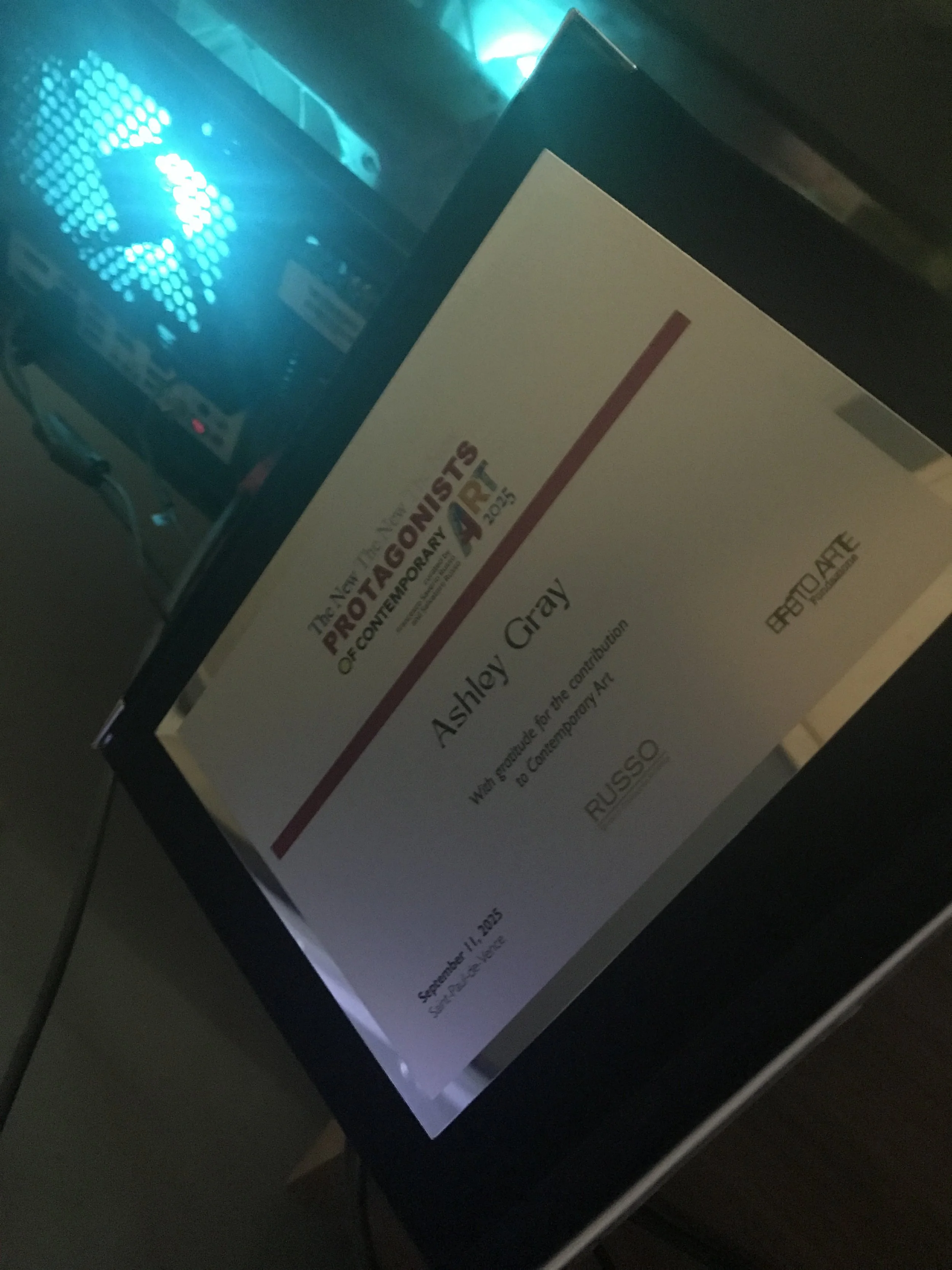 An award certificate titled 'The 13th Still Point of Contemporary Art' awarded to Ashley Cray at RISSO during the event on September 11, 2015, with a blue-lit electronic device in the background.