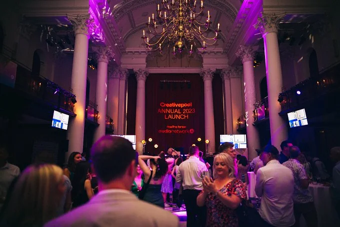 People dancing and socializing in a grand ballroom with high ceilings, large columns, and a chandelier, during a Creativepool Annual 2023 Launch event with purple lighting.
