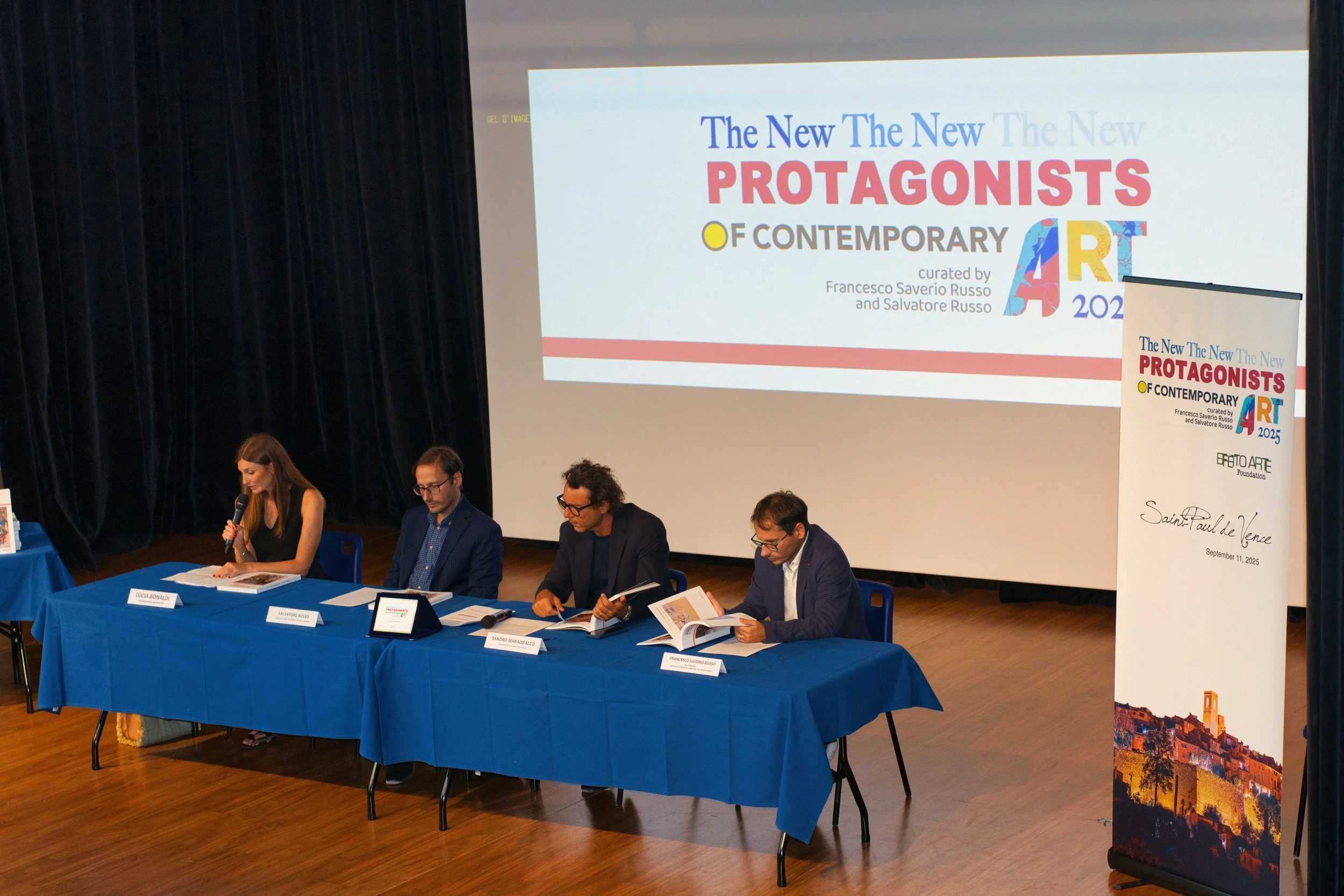 A panel of four people seated at a blue table on stage during a conference. There is a large screen behind them displaying a presentation titled 'The New The New PROTAGONISTS OF CONTEMPORARY ART 2025'. A standing banner to the right also has the title and additional text.