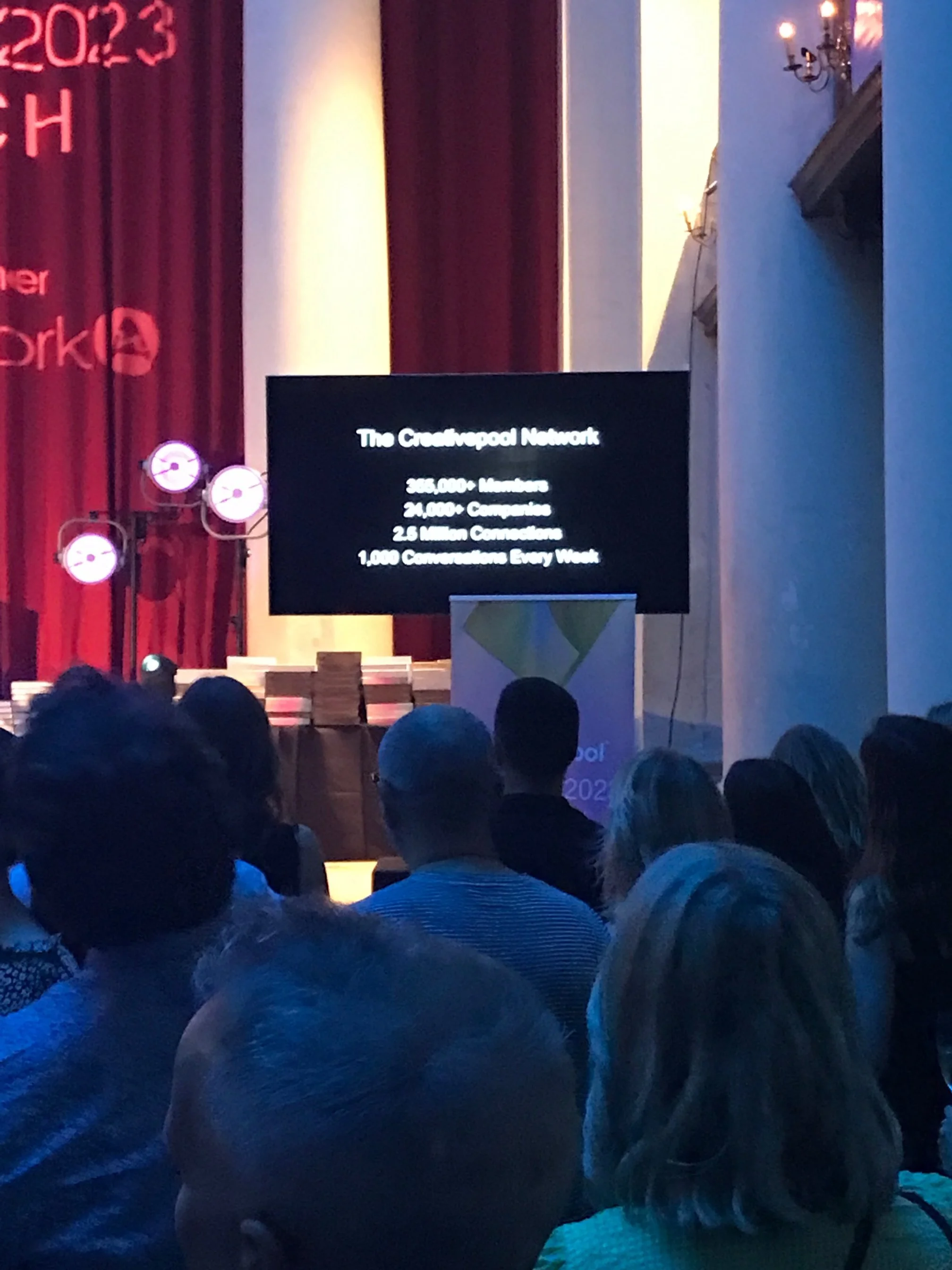 Audience attending a presentation on The Connected Network with a screen showing statistics about members, companies, connections, and weekly conversations, in a conference setting with stage lighting and a large screen.