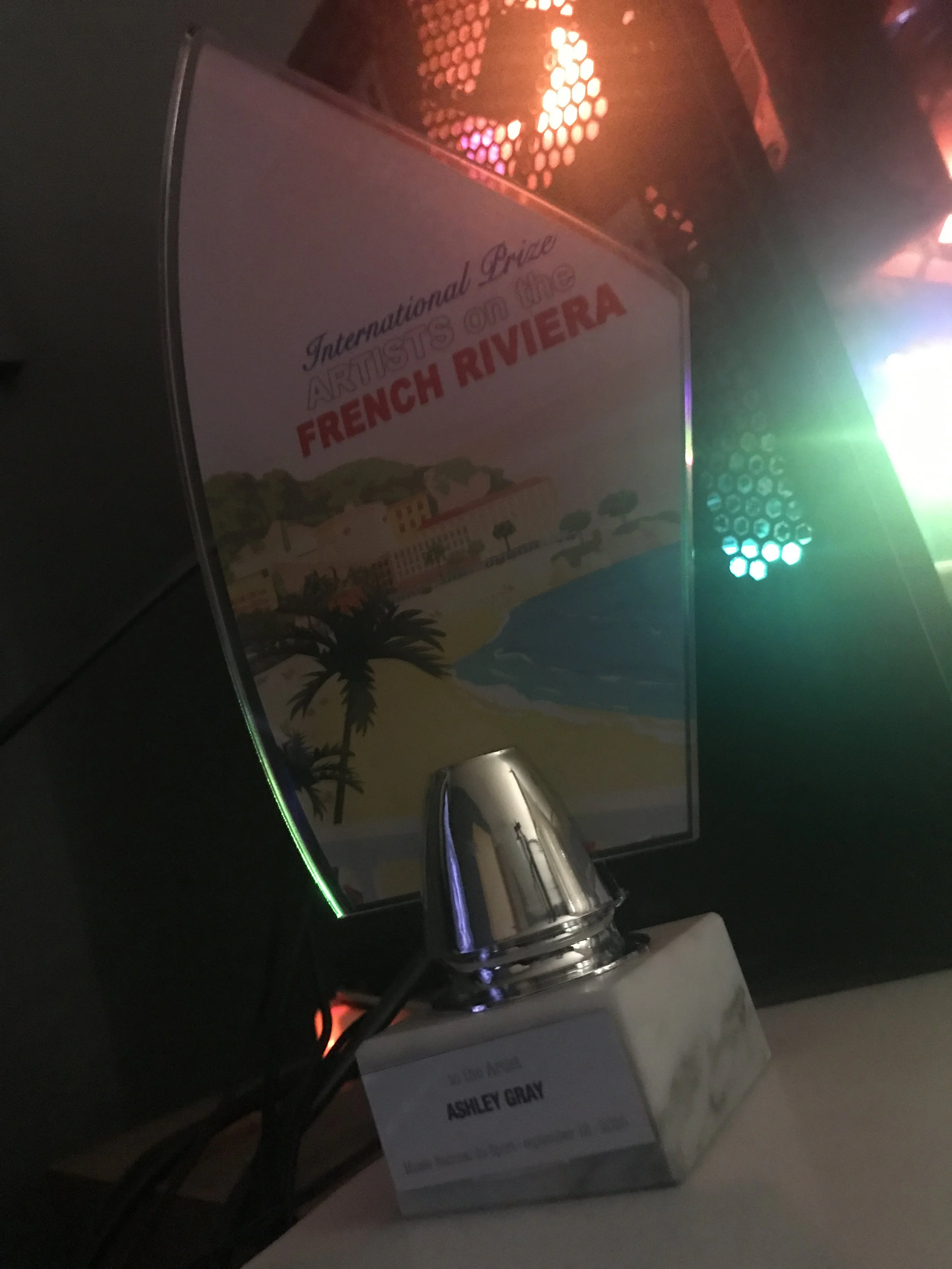 Sign for the International Pride Artists on the Art in the French Riviera exhibit, with a colorful beach scene illustration, a silver bell, and a name tag for Ashley Gray.