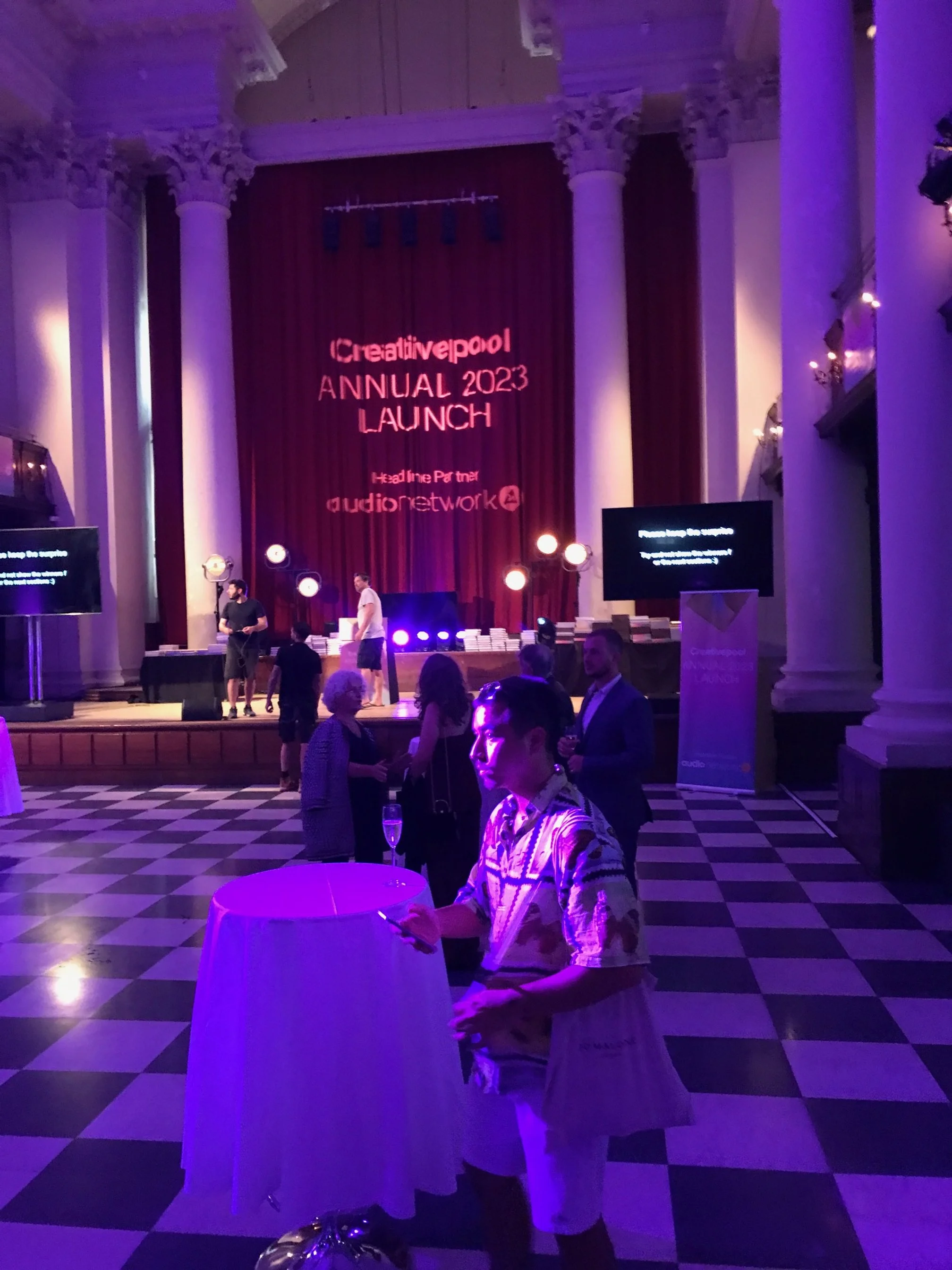 People attending the Creativepool Annual 2023 Launch event in a grand hall with high ceilings and large columns, purple lighting, and a stage with a large red curtain displaying the event name. Several individuals are seen socializing, some standing and some seated, with tables and screens around.