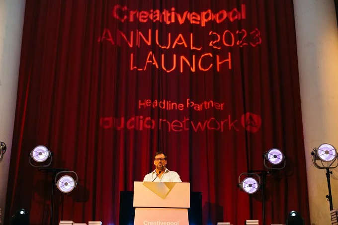 A man standing at a podium on stage during the Creativepool Annual 2023 Launch event, with red curtains and stage lights, and the event name projected on the curtain behind him.