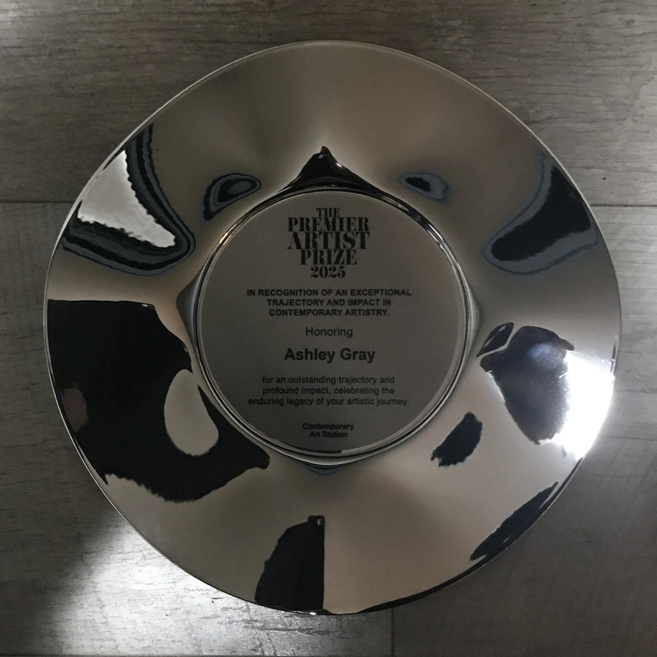 Round shiny silver award plate with engraved text honoring Ashley Gray for outstanding artistic impact, with a reflection of the surroundings.