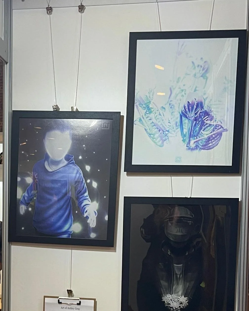The image shows three framed pieces of artwork hanging on a white wall in an art gallery. The left piece features a person in a hoodie with a blank face, surrounded by glowing orbs and light effects. The top right piece depicts abstract, colorful shapes resembling a stylized bouquet or flowers. The bottom right piece appears to be a dark portrait or abstract figure with a reflective or glossy appearance.