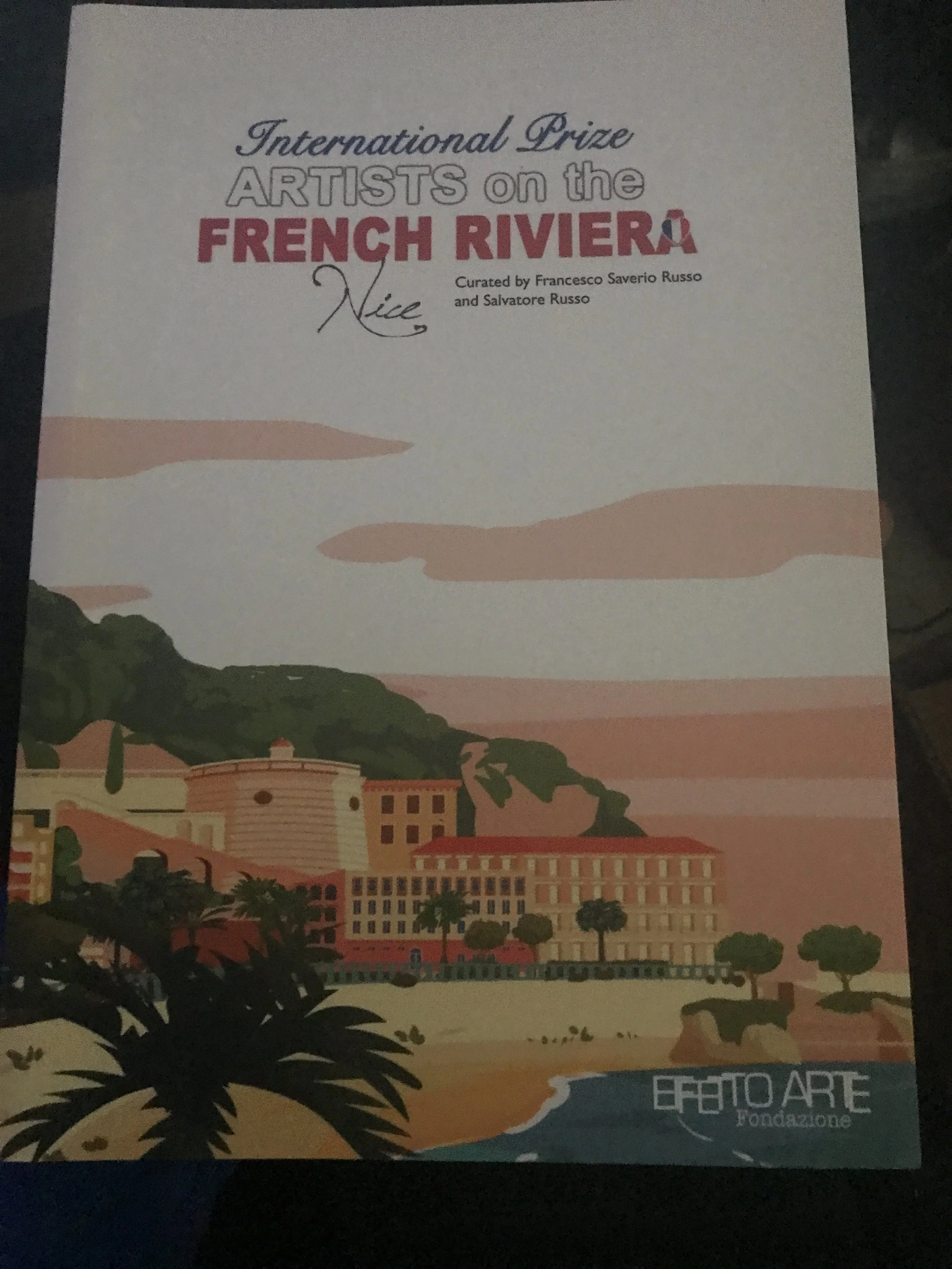 Cover art for an art exhibition titled "Artists on the French Riviera" curated by Francesco Saverio Russo and Salvatore Russo, featuring a stylized illustration of a hillside town with buildings, trees, and a coastline, with a soft pastel color palette.