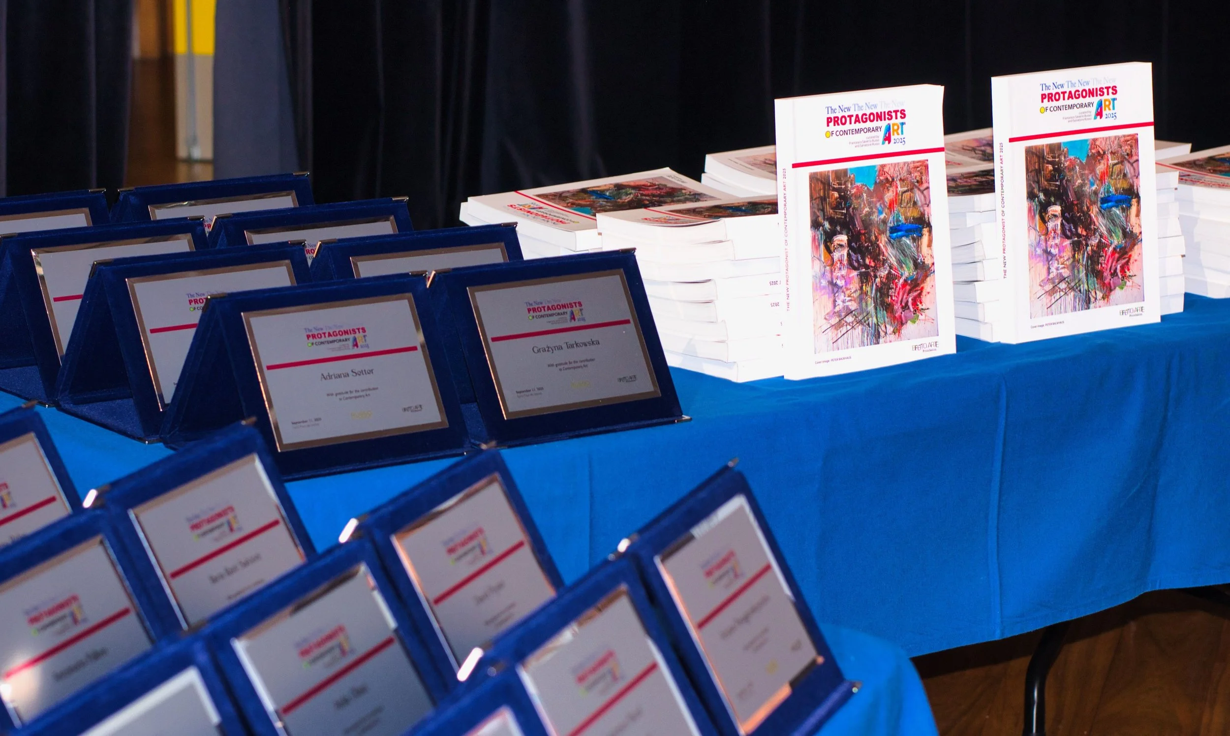 Table with awards certificates and copies of an art book titled 'The New Protagonists of Contemporary Art' featuring a colorful abstract painting on the cover.