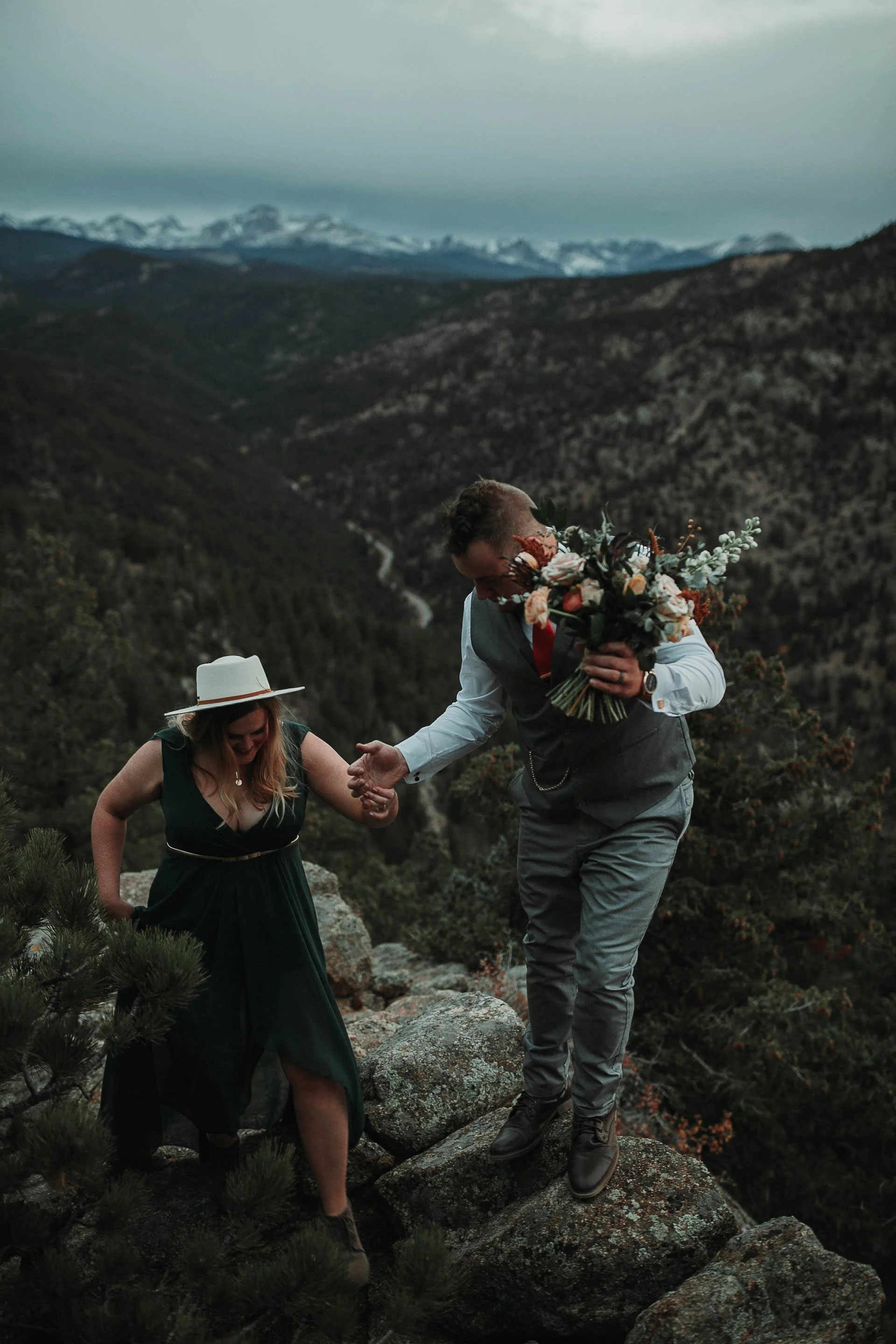 Rocky Mountain Elopement photo of groom helping bride climb up the  cliffside with the snowcap mountains behind them