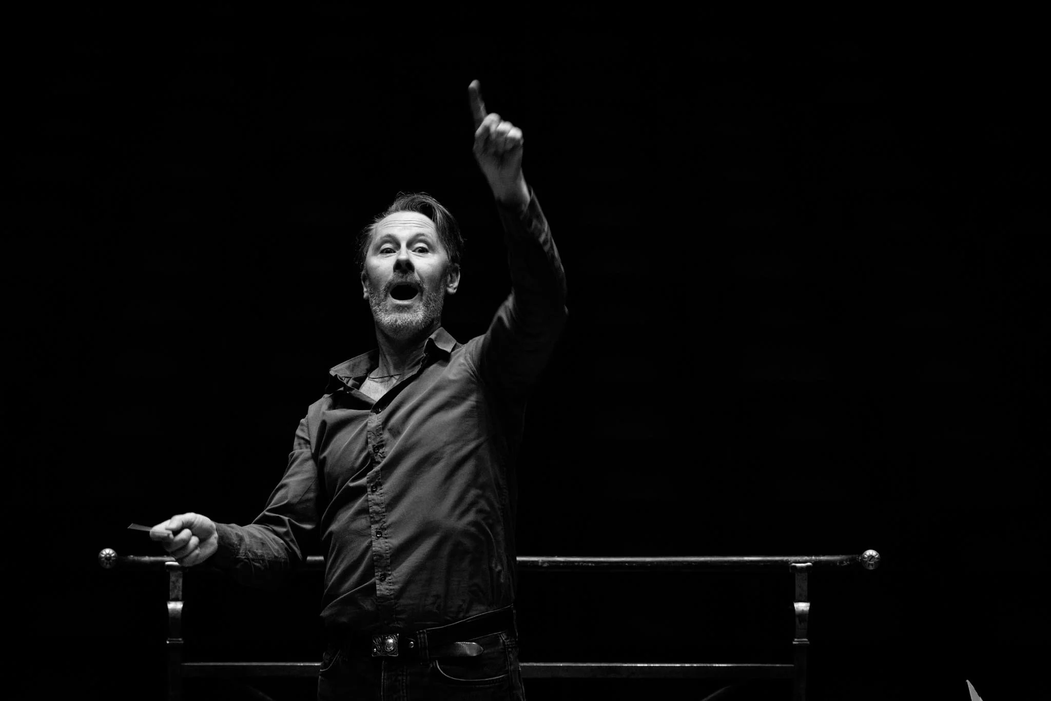 A man with a beard and dressed in dark clothing is pointing upward with his right hand conduction, standing behind a rail on a dark stage.