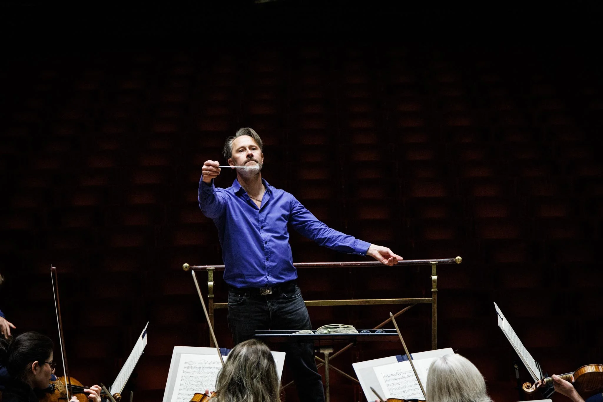 A man in a blue shirt conducting an orchestra in a dark concert hall.