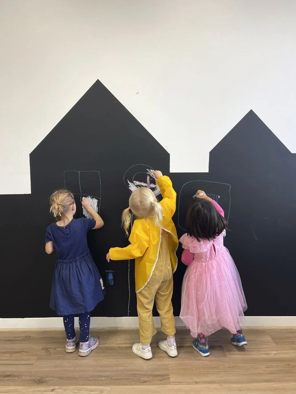 Three children drawing on a black chalkboard wall with chalk, with house shapes sketched on the upper part of the wall.