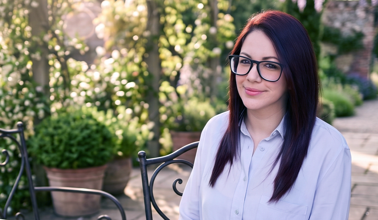 A woman with long dark hair and glasses sitting outside on a patio surrounded by greenery and potted plants, smiling softly.