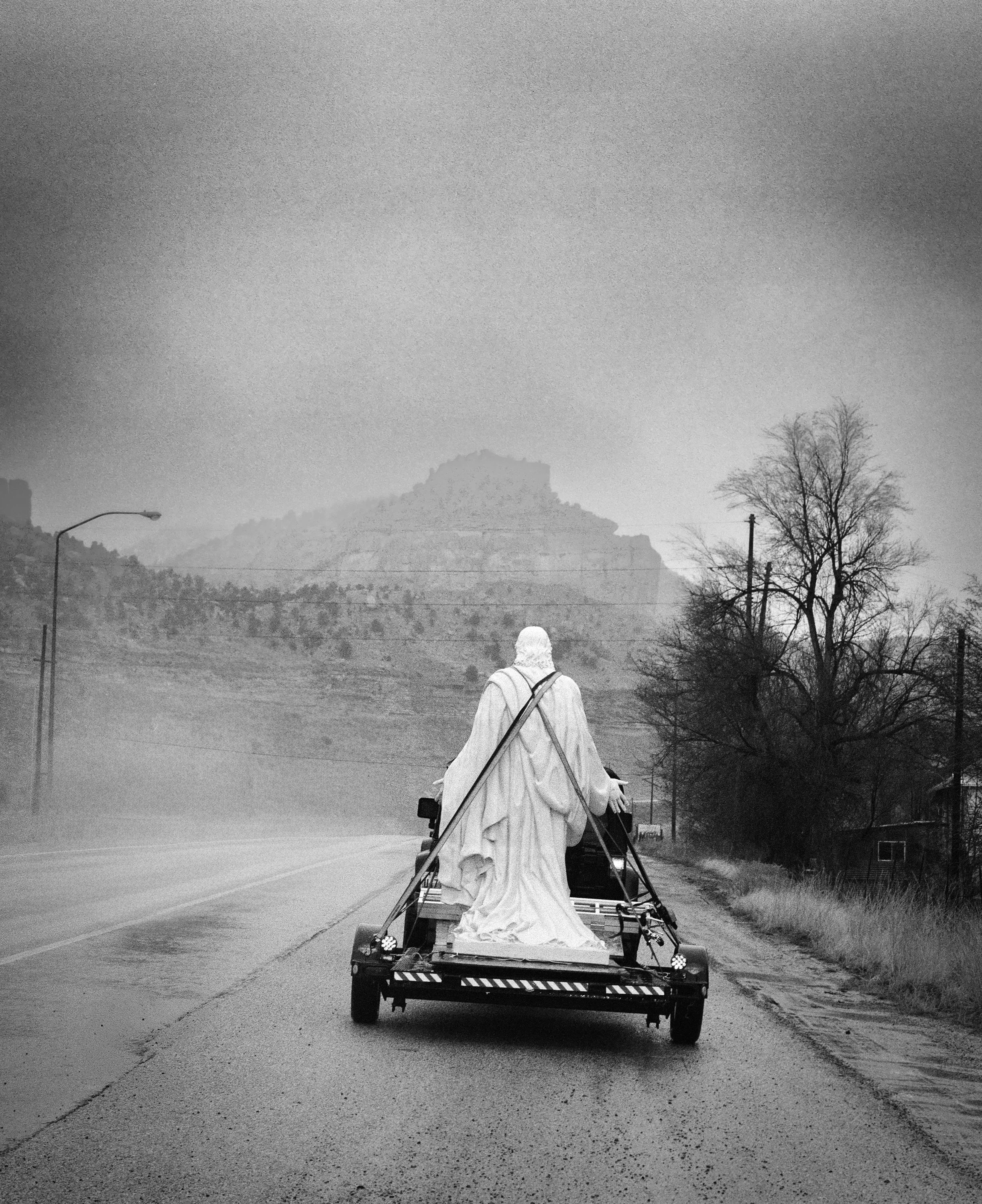 A black and white medium format film photograph of a marble statue of Jesus strapped onto a flatbed trailer driving down a misty highway in the American West.