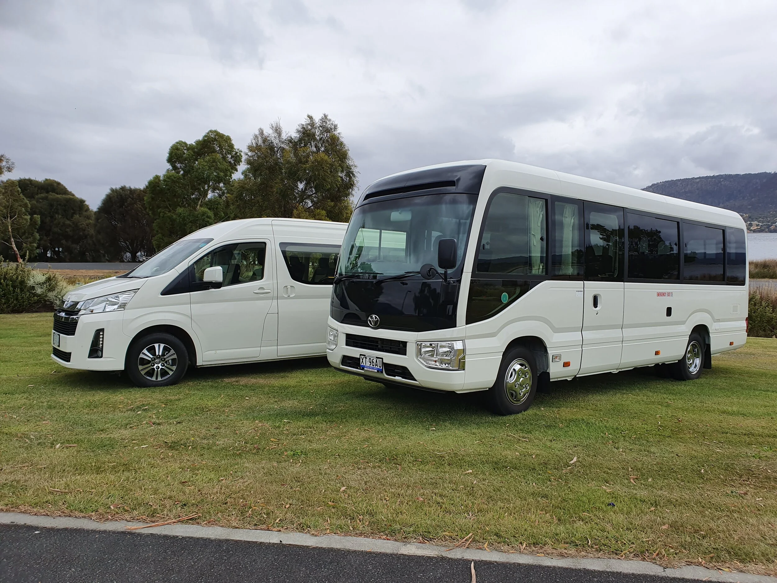 Two white vans parked on grass with trees in the background.