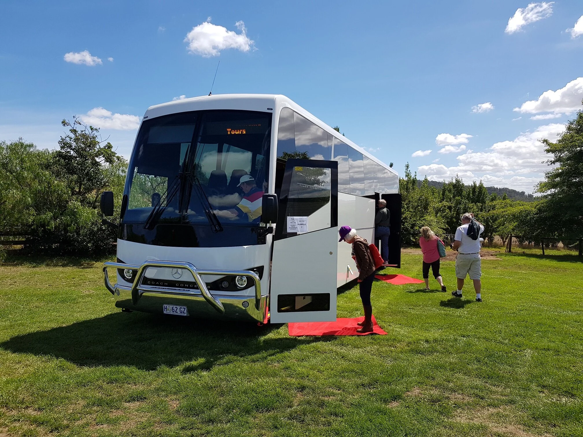 A tour bus parked on a grassy area with passengers boarding. The bus is white and labeled "Tours," with an open door and a red carpet leading inside. Several people are seen walking towards and entering the bus. The background includes trees and a br