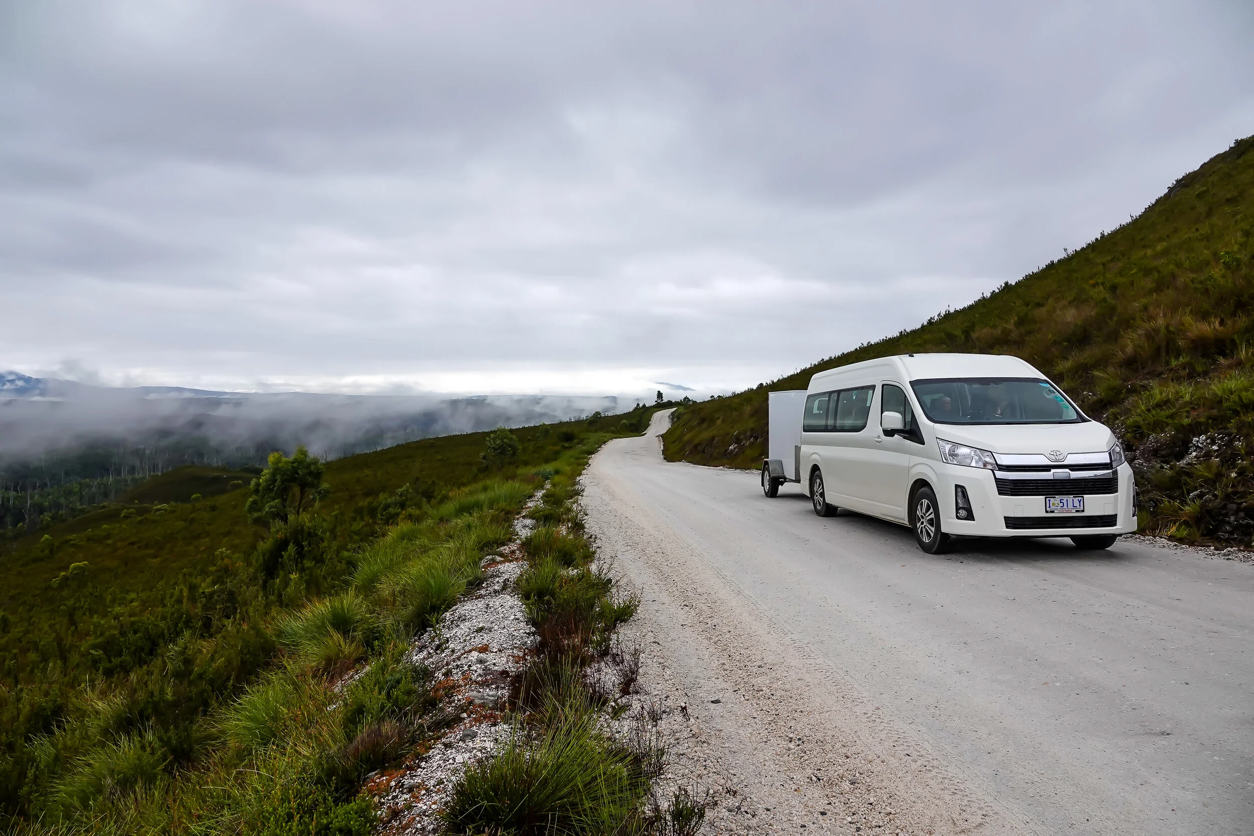 White van with a trailer on a remote winding road surrounded by hills and greenery, under a cloudy sky.