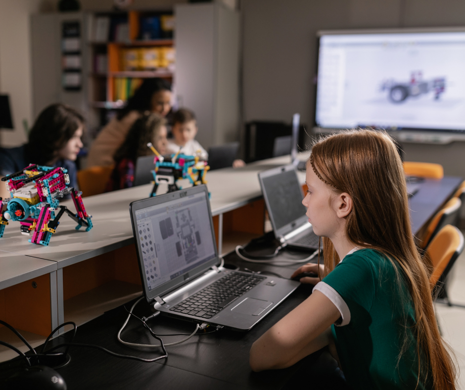 A young girl working on a laptop in a classroom setting with robotics models on the desk. A group of people, including children, is visible in the background, engaging with technology. A screen displays a 3D model of a robot.