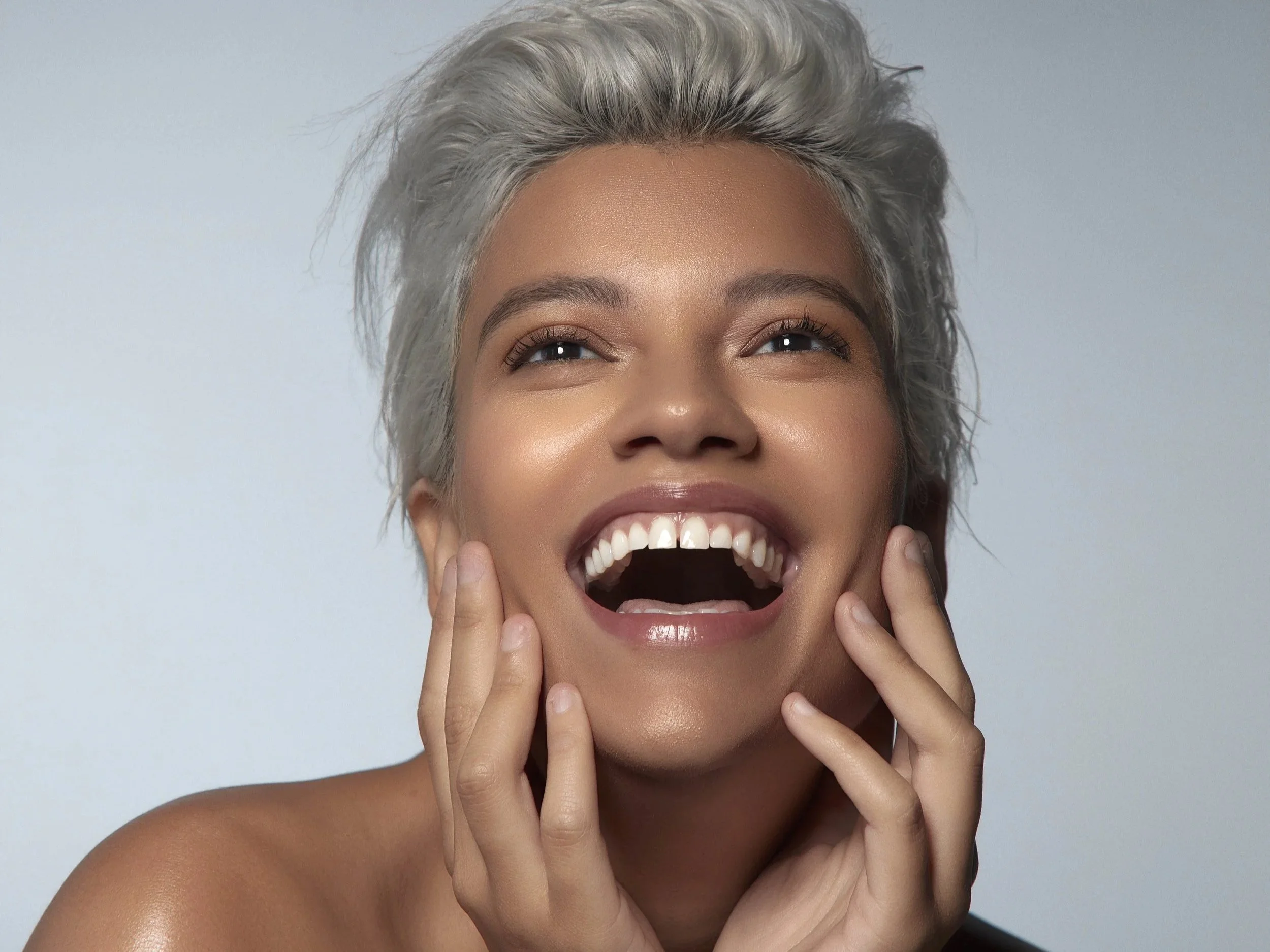 A woman with short, grey hair smiling with her mouth open, showing her teeth, and touching her face with her hands, against a plain light background.