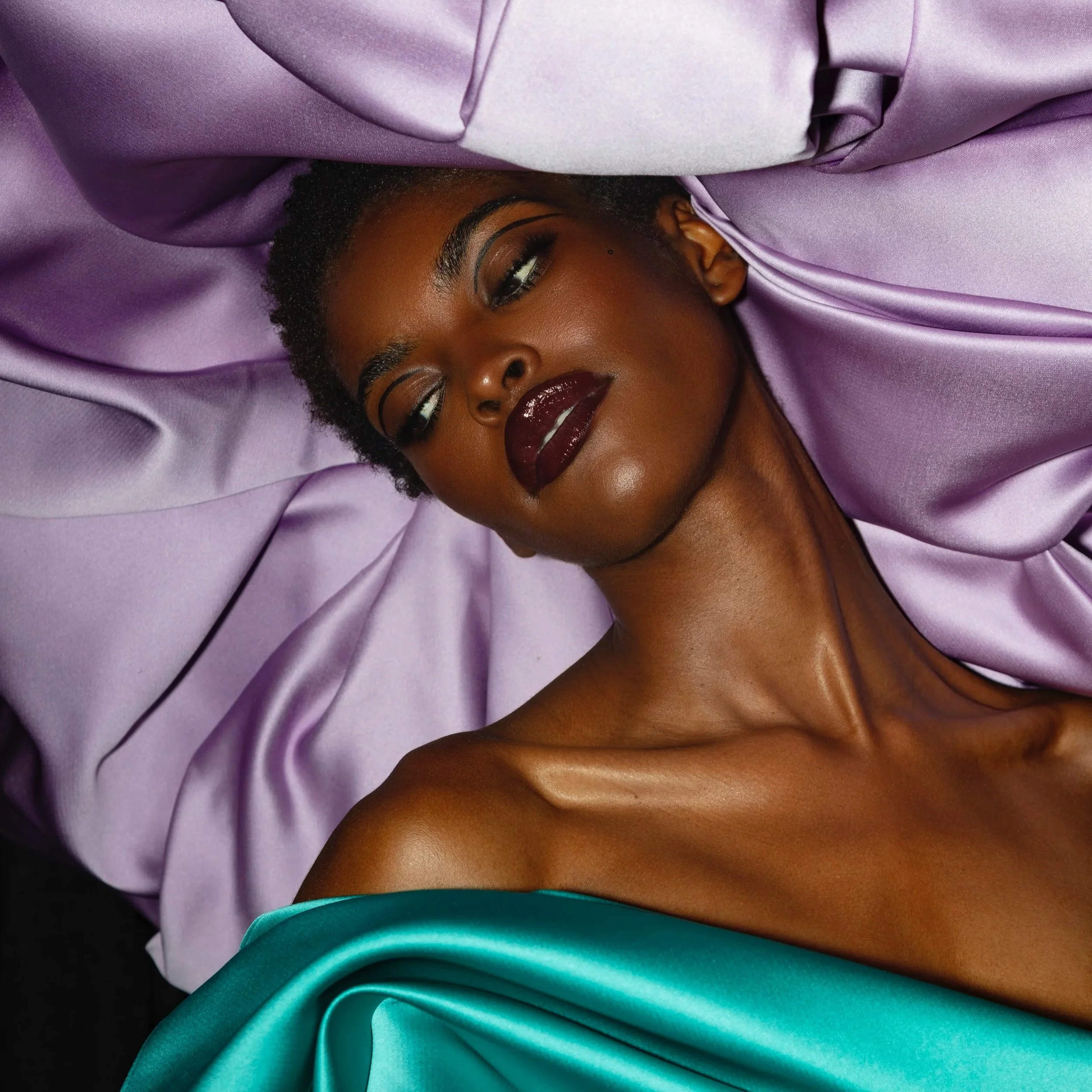 A woman with dark lipstick and makeup, lying on purple satin fabric with a teal satin garment partially covering her shoulder.