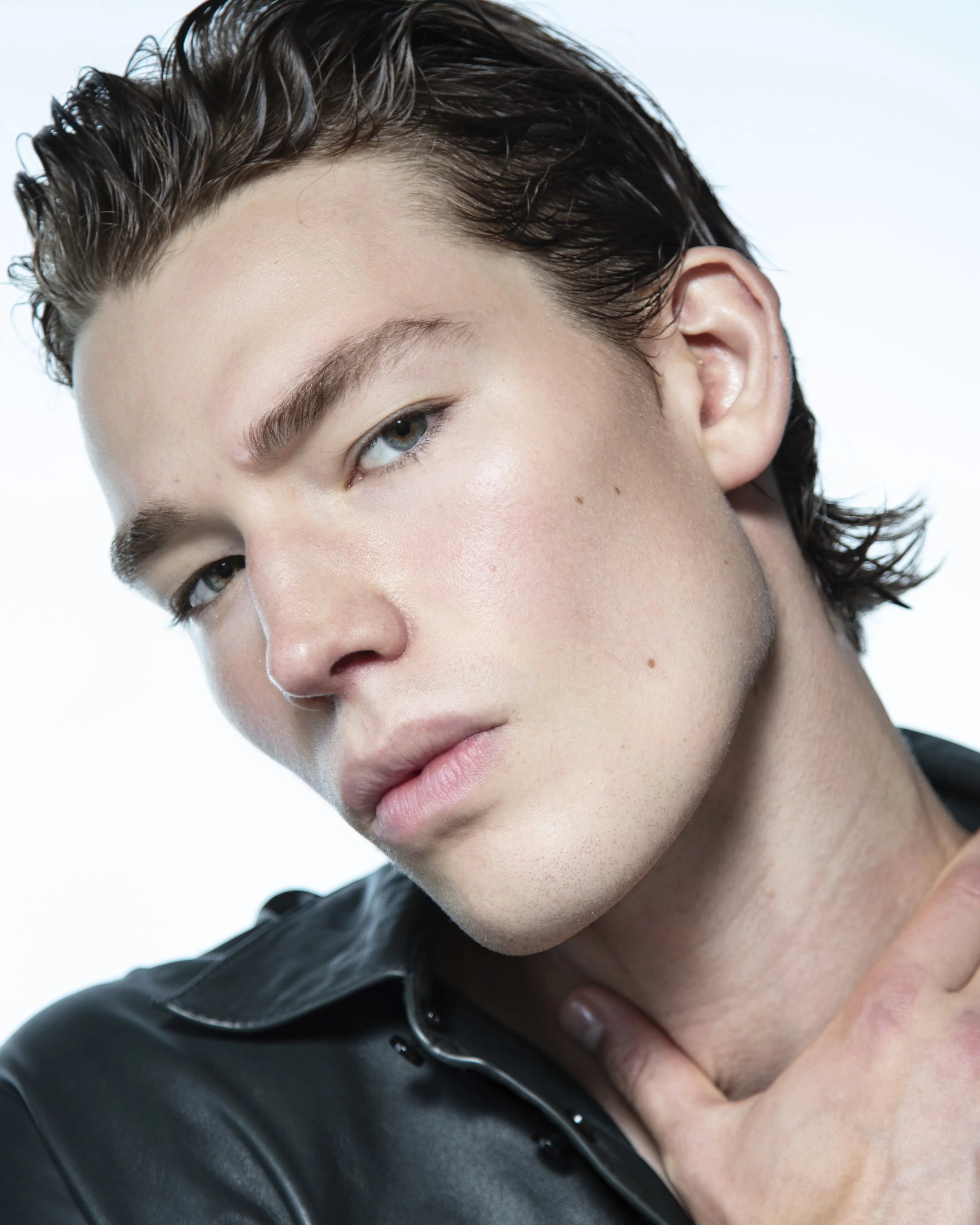 Close-up portrait of a young man with wet, styled hair, piercing blue eyes, wearing a black leather jacket, touching his neck with one hand.